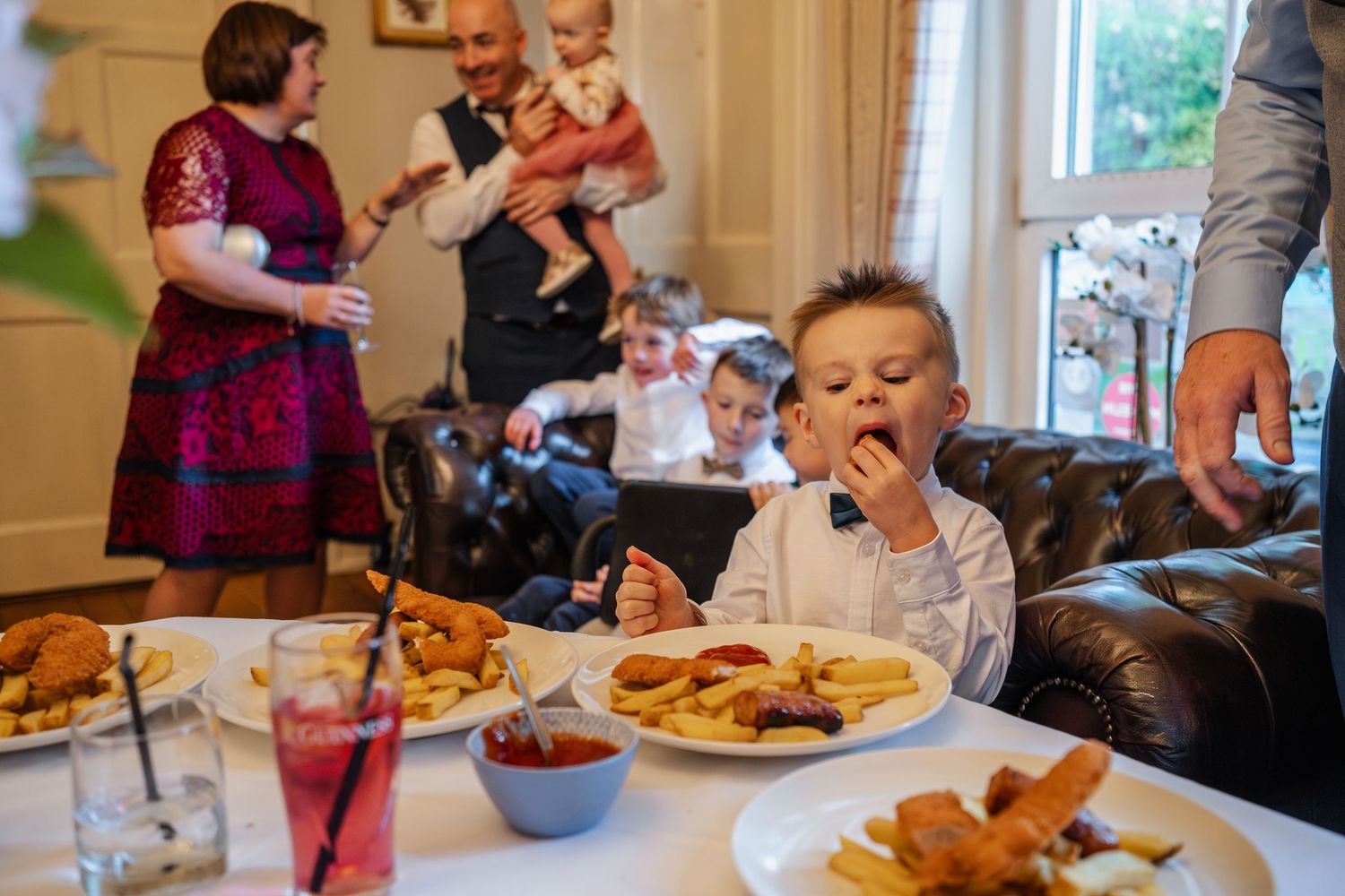 Young boy in bow tie eating at a wedding reception table with family gathered in the background.