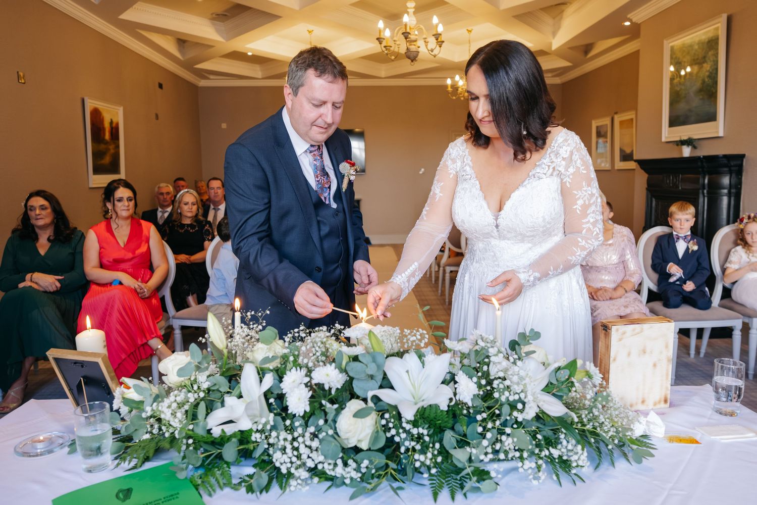 Bride and groom lighting unity candle at wedding ceremony altar decorated with white flowers and greenery.