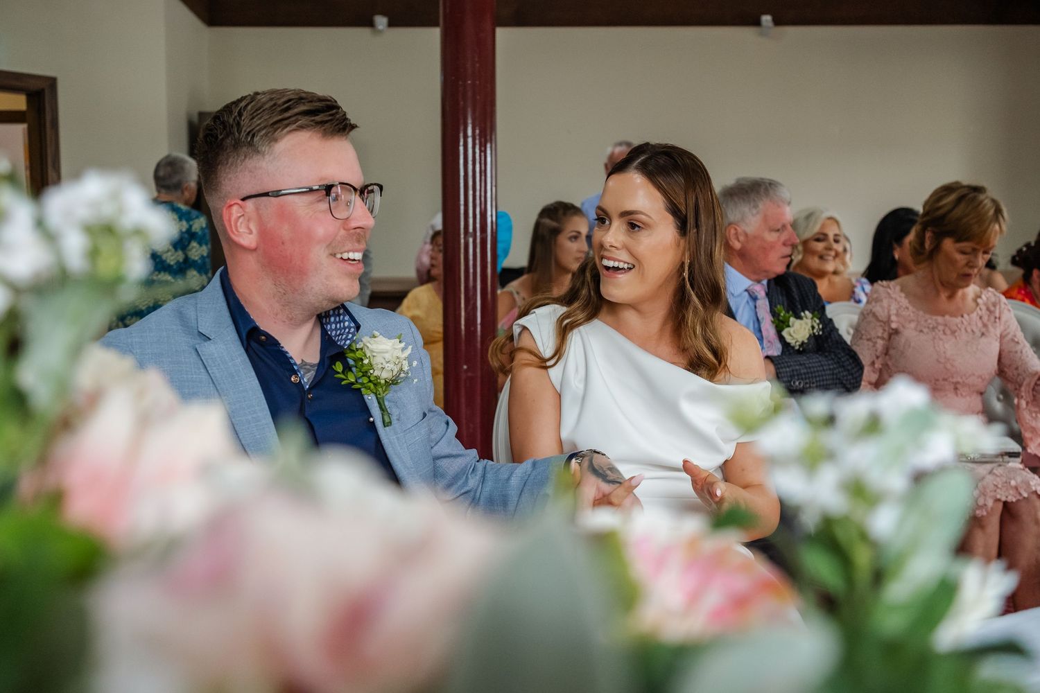 Bride and groom laughing together at wedding ceremony, surrounded by pink and white floral arrangements.
