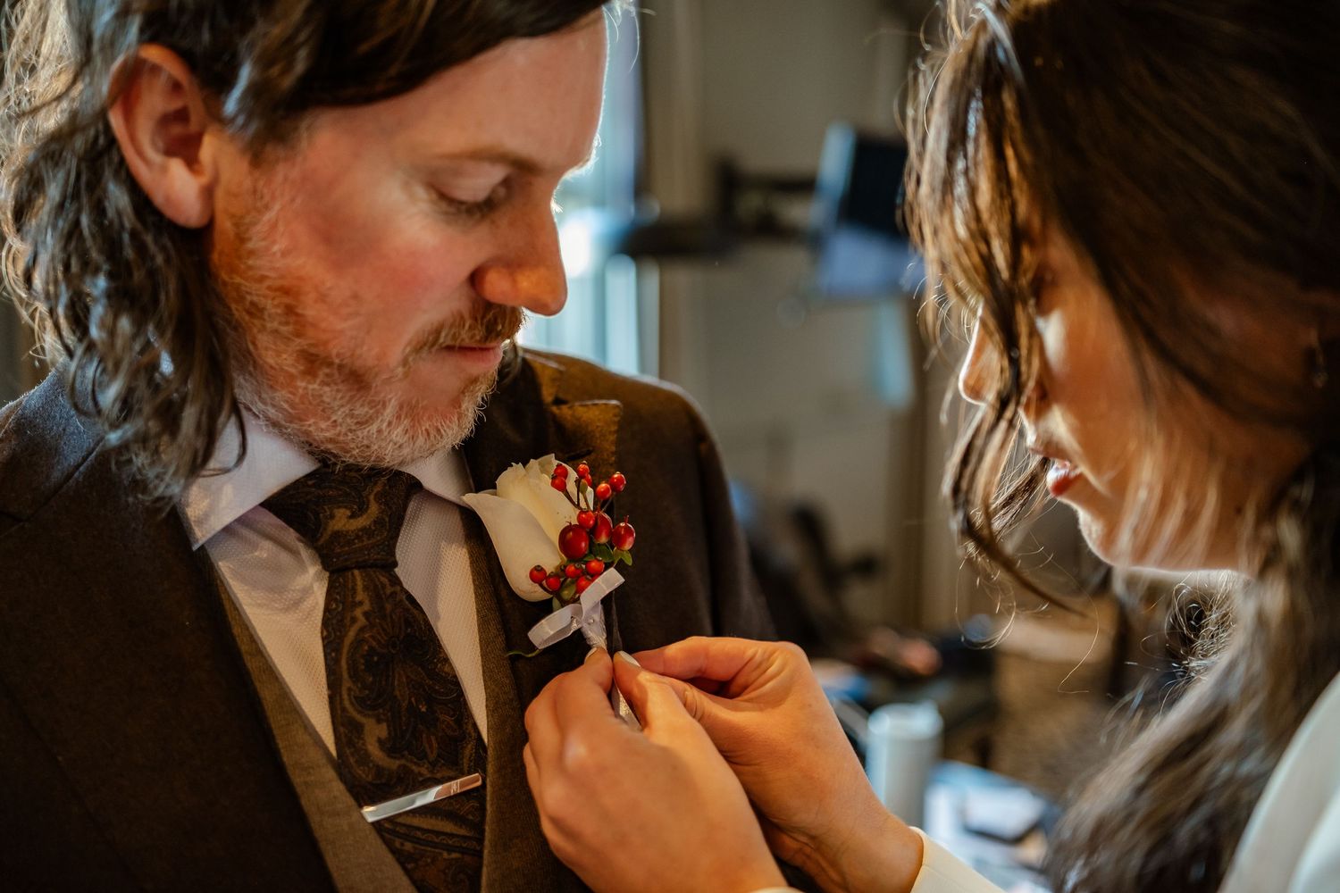 A woman pins a floral boutonniere onto a mans brown suit jacket during wedding preparations.