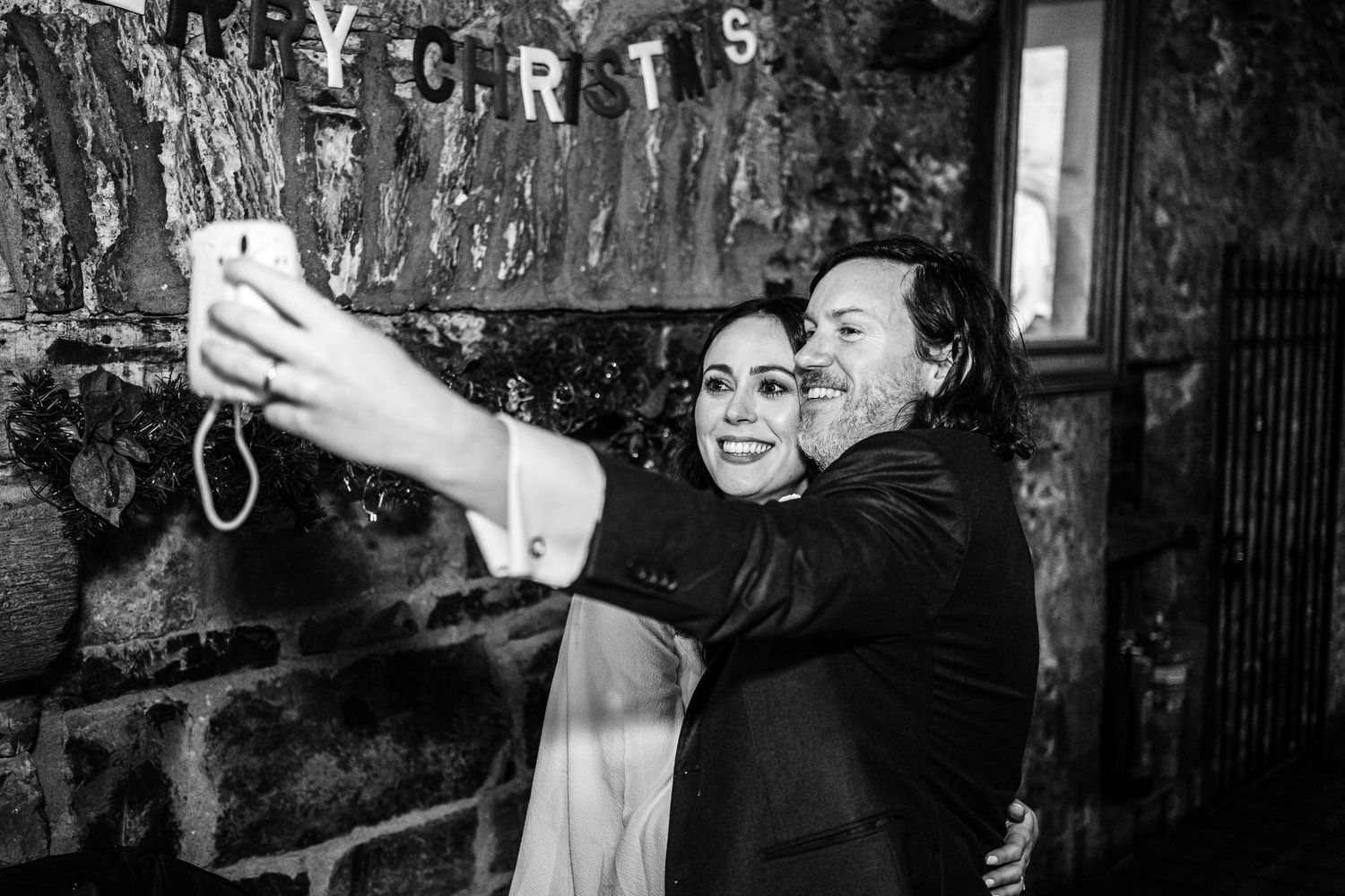 Bride and groom taking a selfie together at their wedding reception in front of a rustic stone wall.