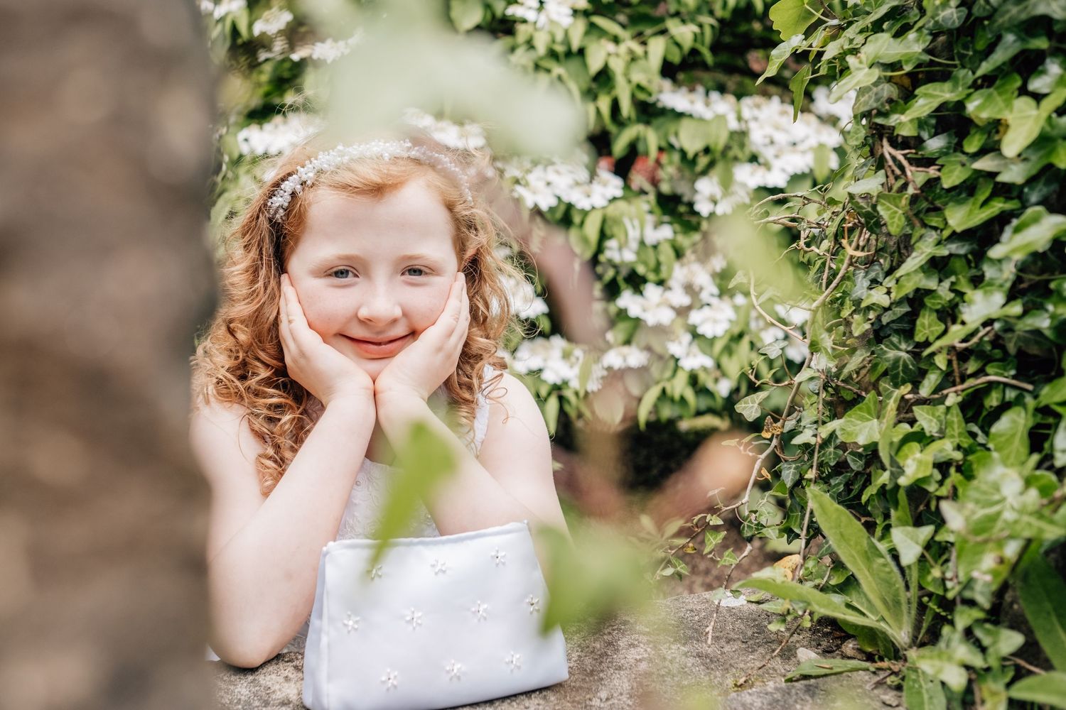 A smiling red-haired girl in a white dress rests her chin on her hands among lush green bushes with white flowers.