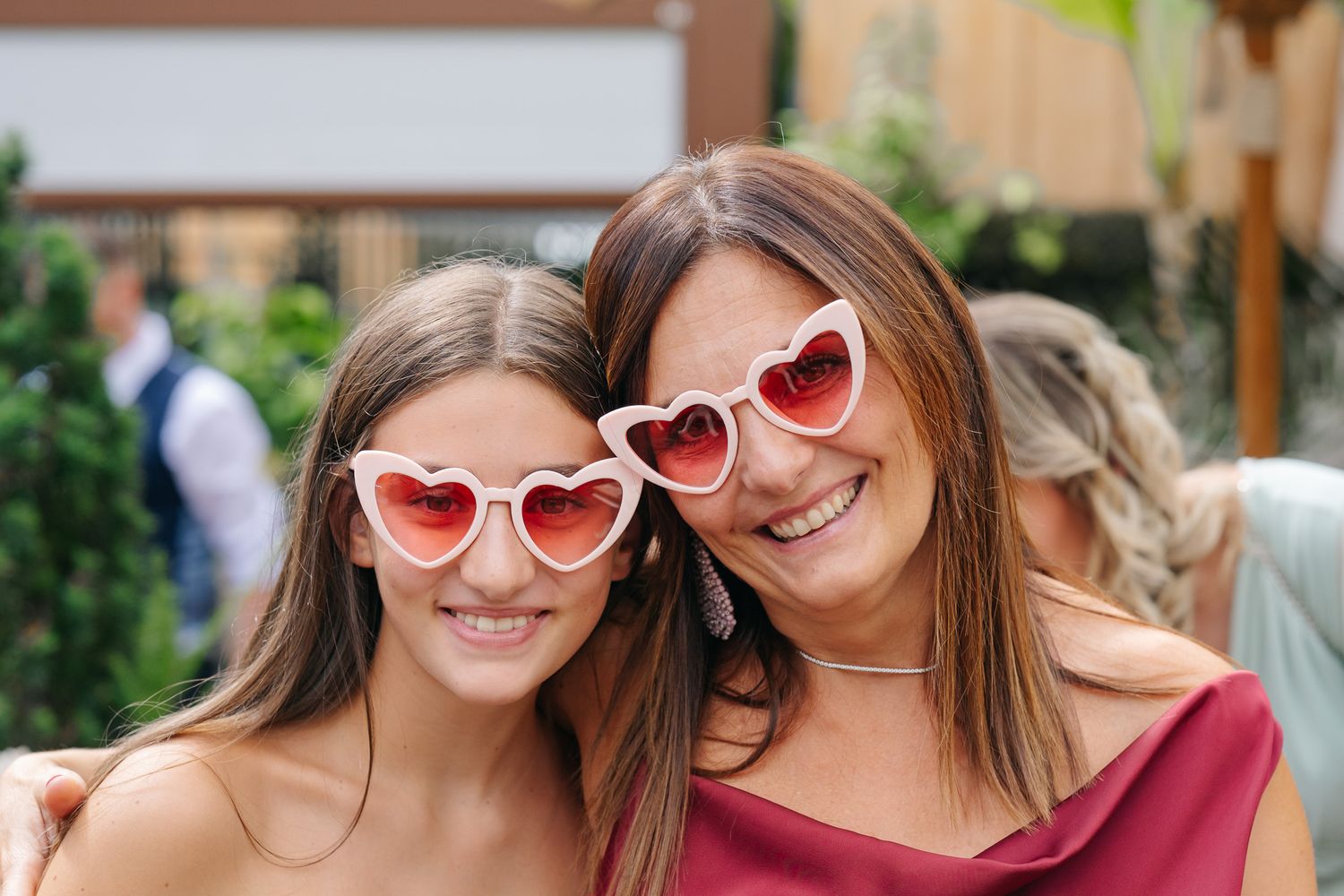 Two women wearing matching heart-shaped pink sunglasses smile together outdoors at a festive event.
