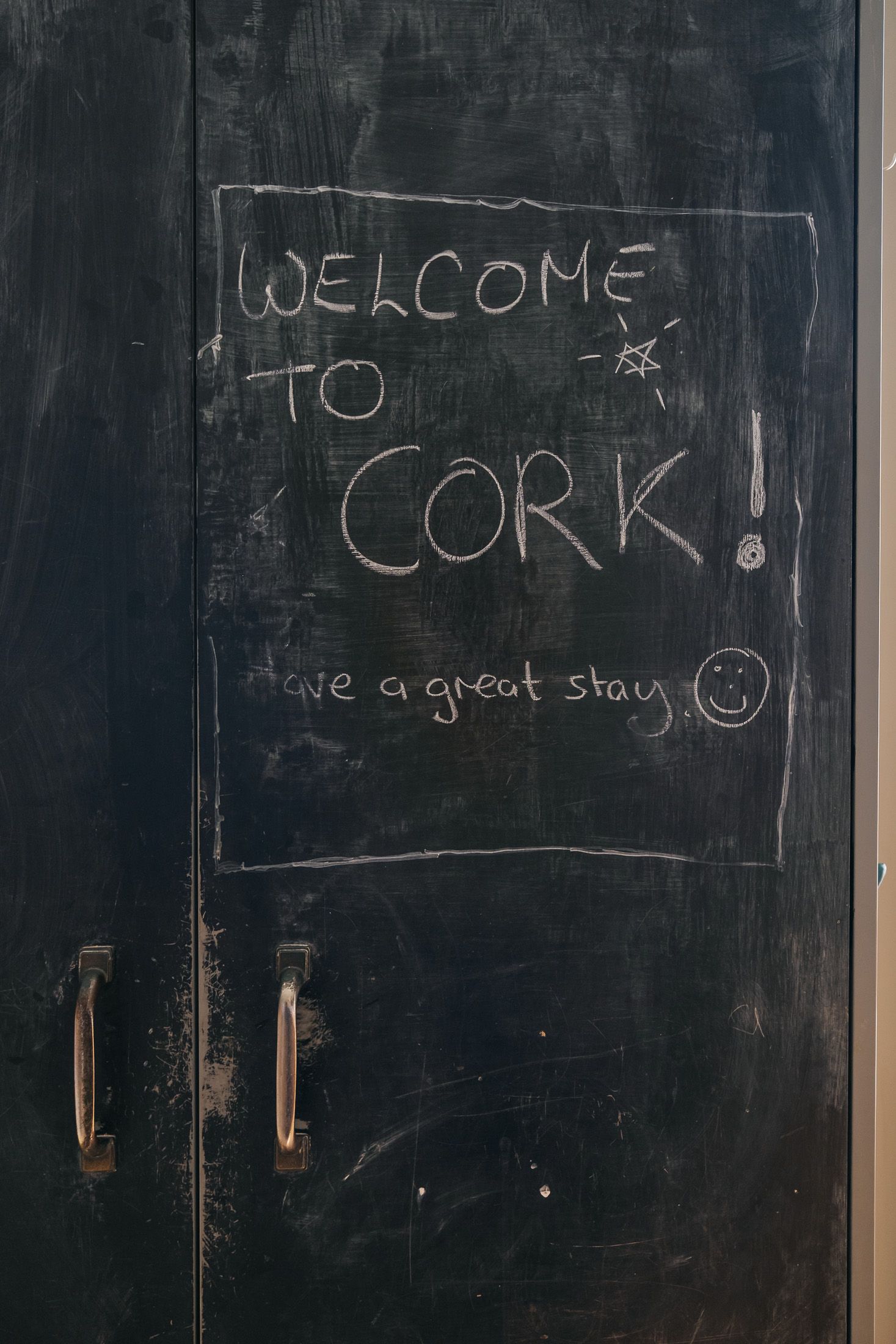 Chalkboard door with 'Welcome to Cork! Have a great stay' written in chalk with a smiley face.