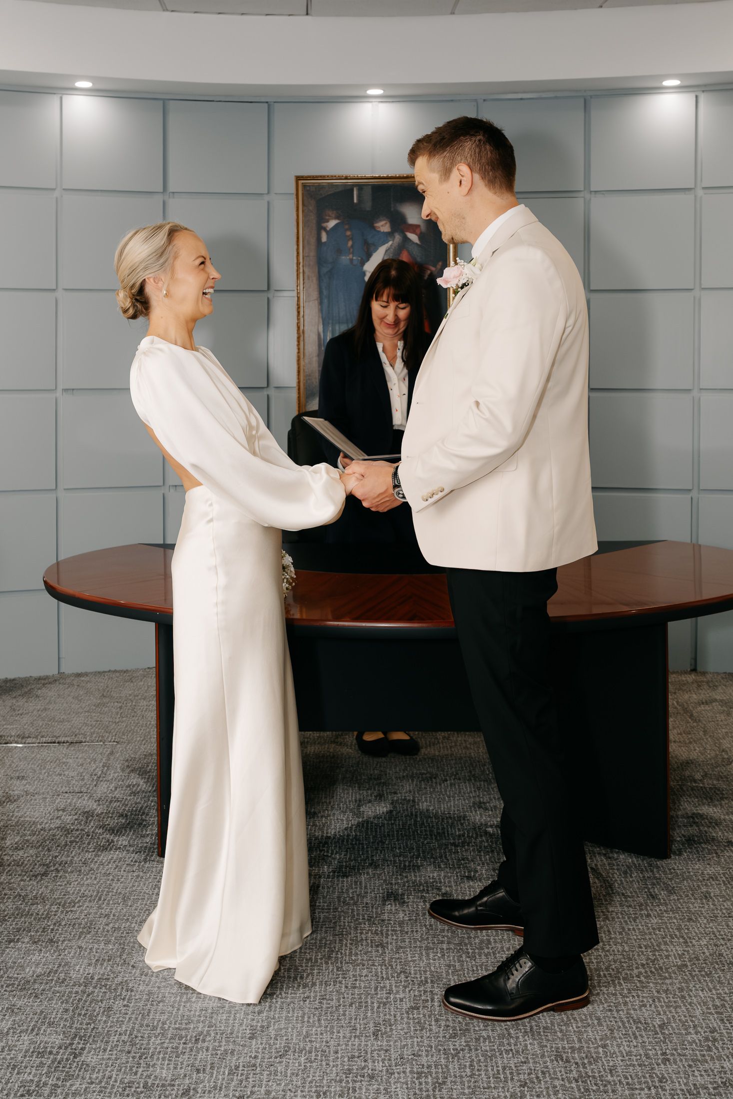 Bride and groom exchange vows at a civil wedding ceremony, holding hands before an officiant at a wooden desk.
