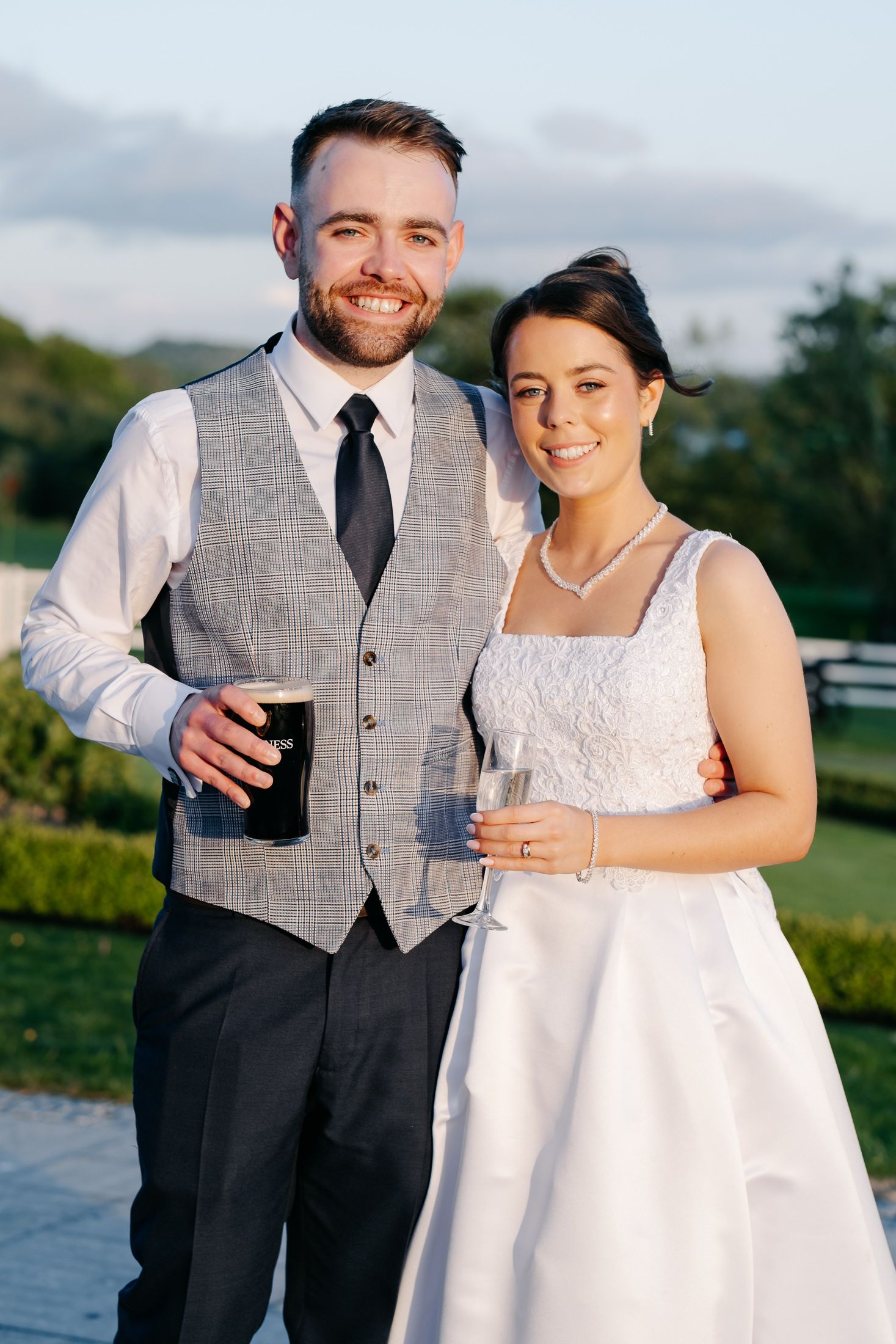 Smiling bride and groom holding drinks outdoors at golden hour, bride in white lace dress, groom in grey vest and black tie.