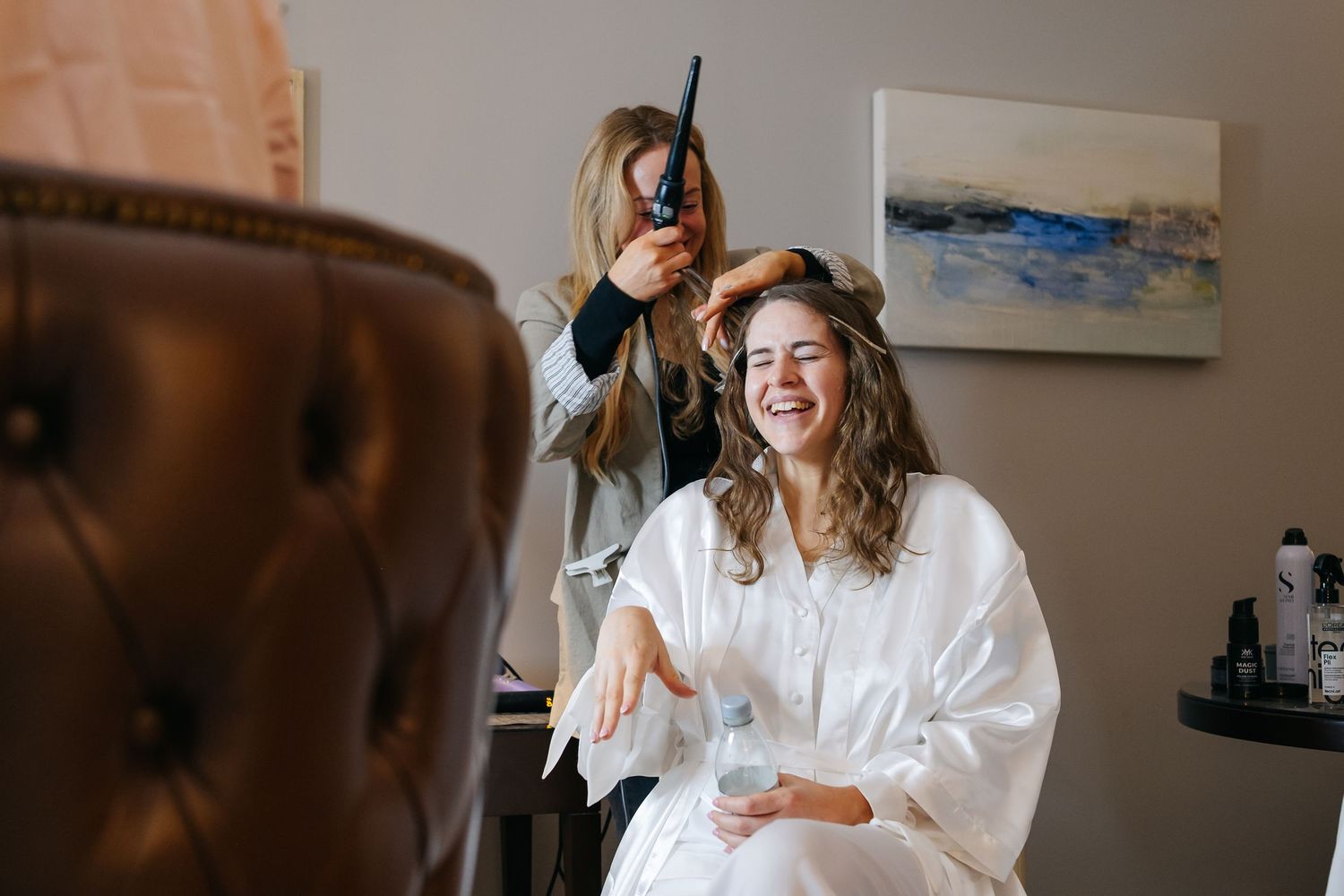 Women in white robes laughing and celebrating together during a bridal preparation moment.