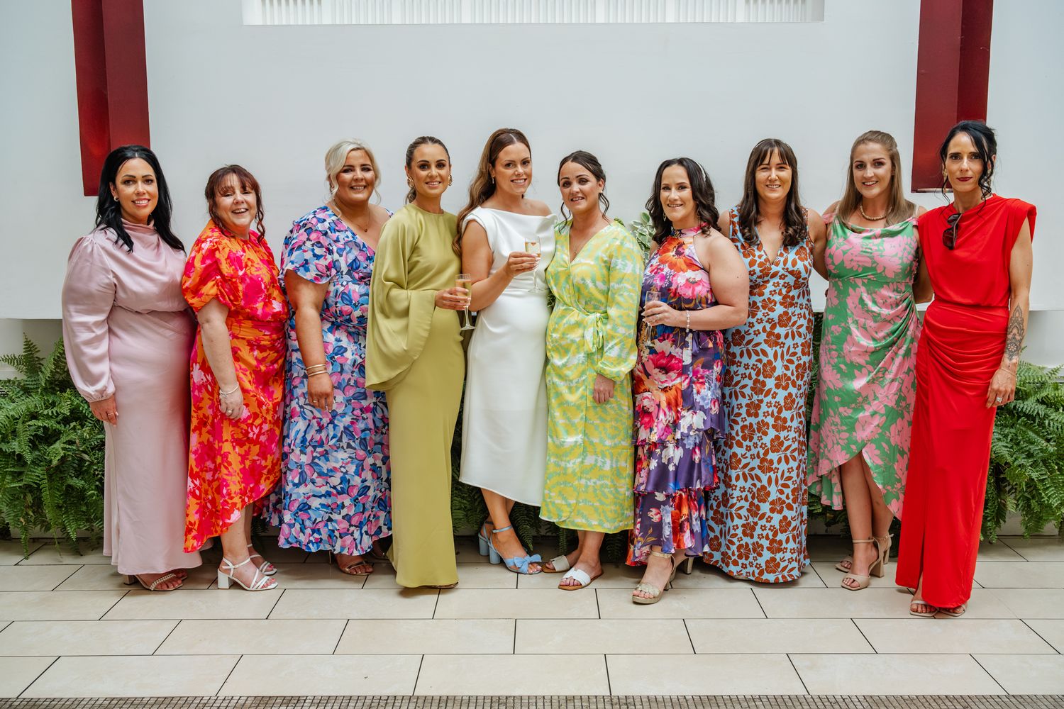 Colorful group photo of wedding party in vibrant dresses against white wall.