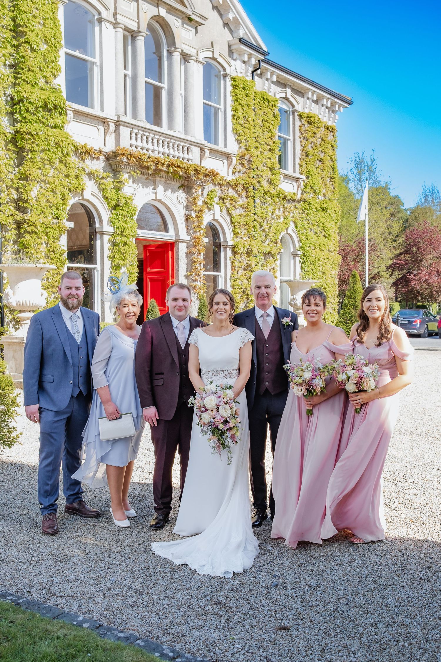 Large formal celebration group poses outdoors at elegant ivy-covered venue with red door under bright blue sky.