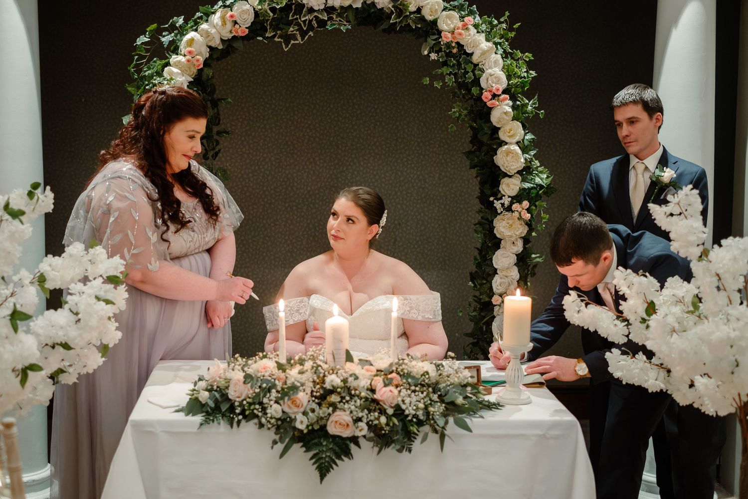 Wedding ceremony under a floral arch with pink roses and white blooms during signing ritual.