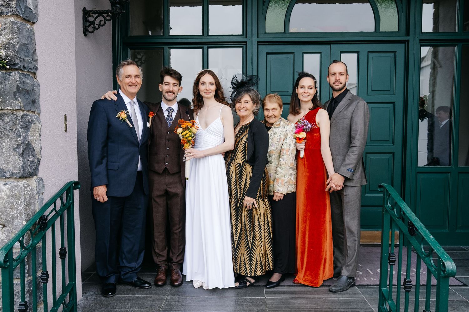 A group of formally dressed wedding guests stand outside a teal-colored doorway of a historic building.