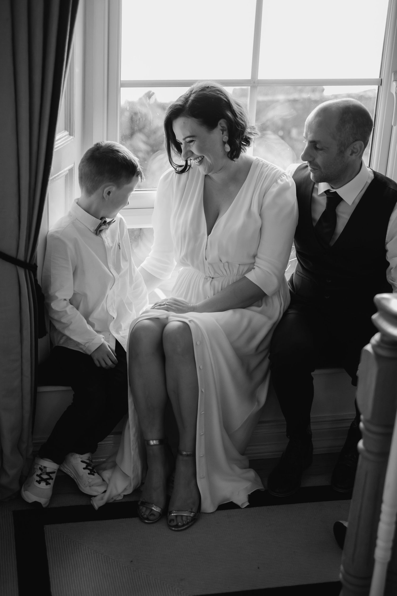 Black and white wedding photo of a bride in white dress laughing with groom and young boy by a bright window.