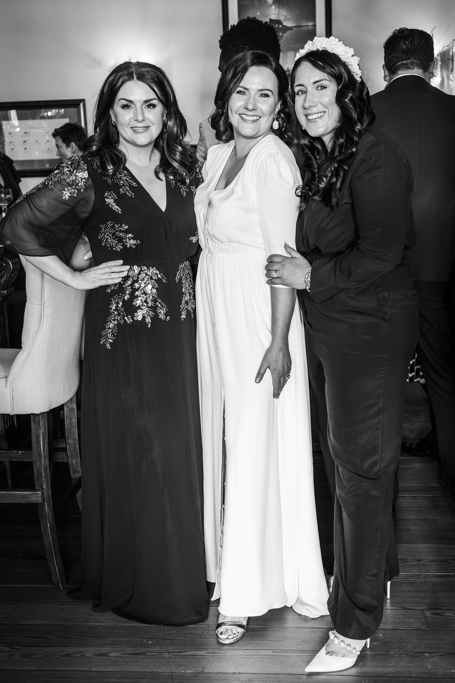 Three women pose together at a wedding, one in a white bridal gown flanked by two guests.
