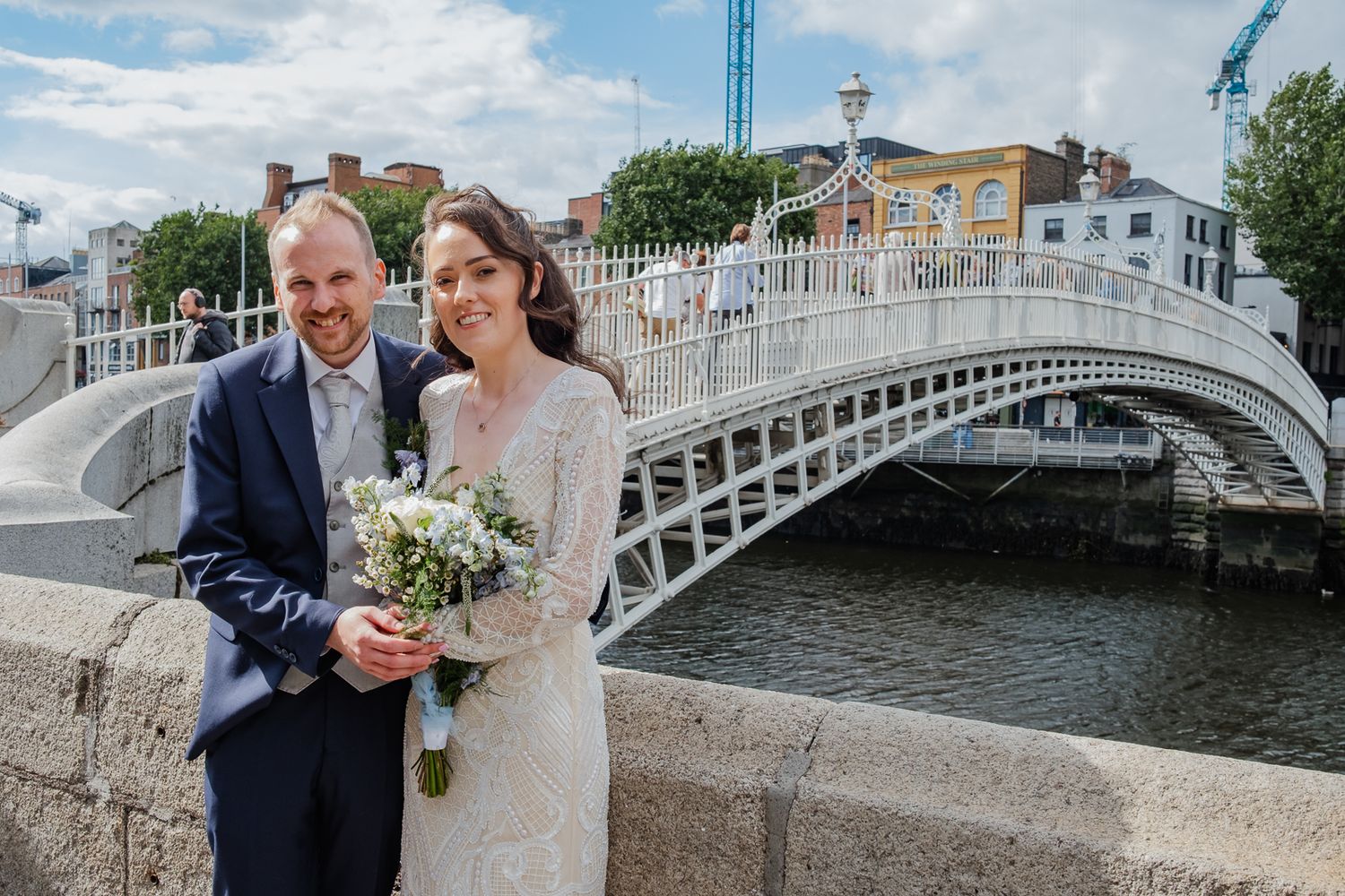Wedding couple posing on a scenic bridge over water with historic architecture in background.