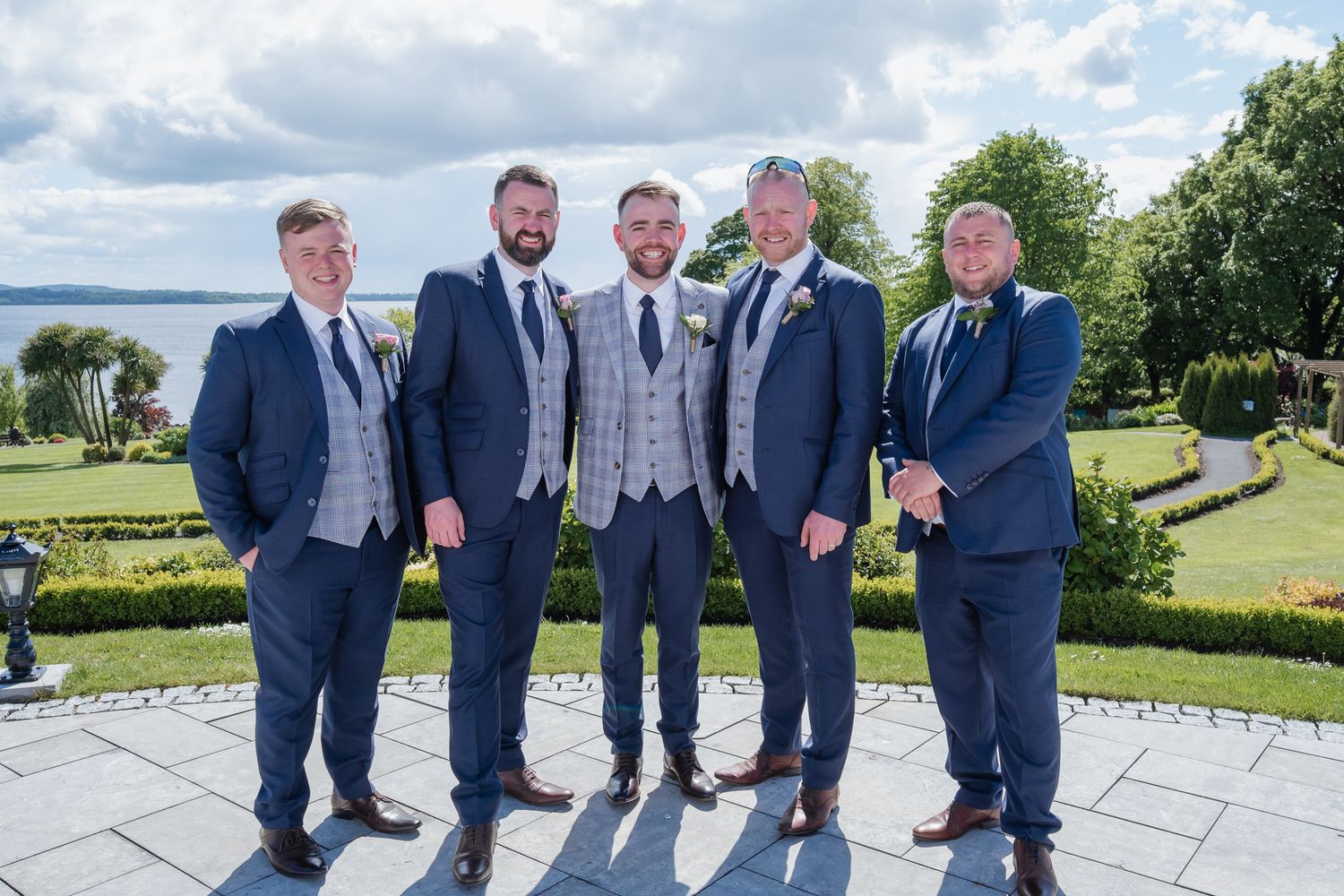 Five groomsmen in navy blue suits posing outdoors at a wedding venue with scenic water views.