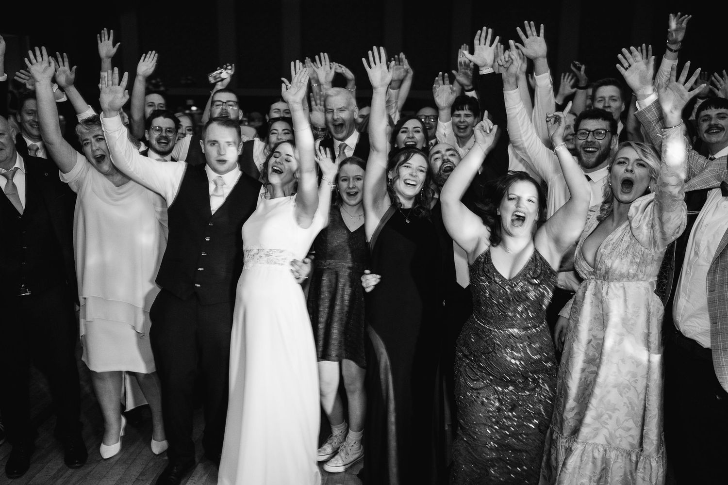 Joyful party crowd raises their hands in celebration at a reception with vibrant purple lighting.