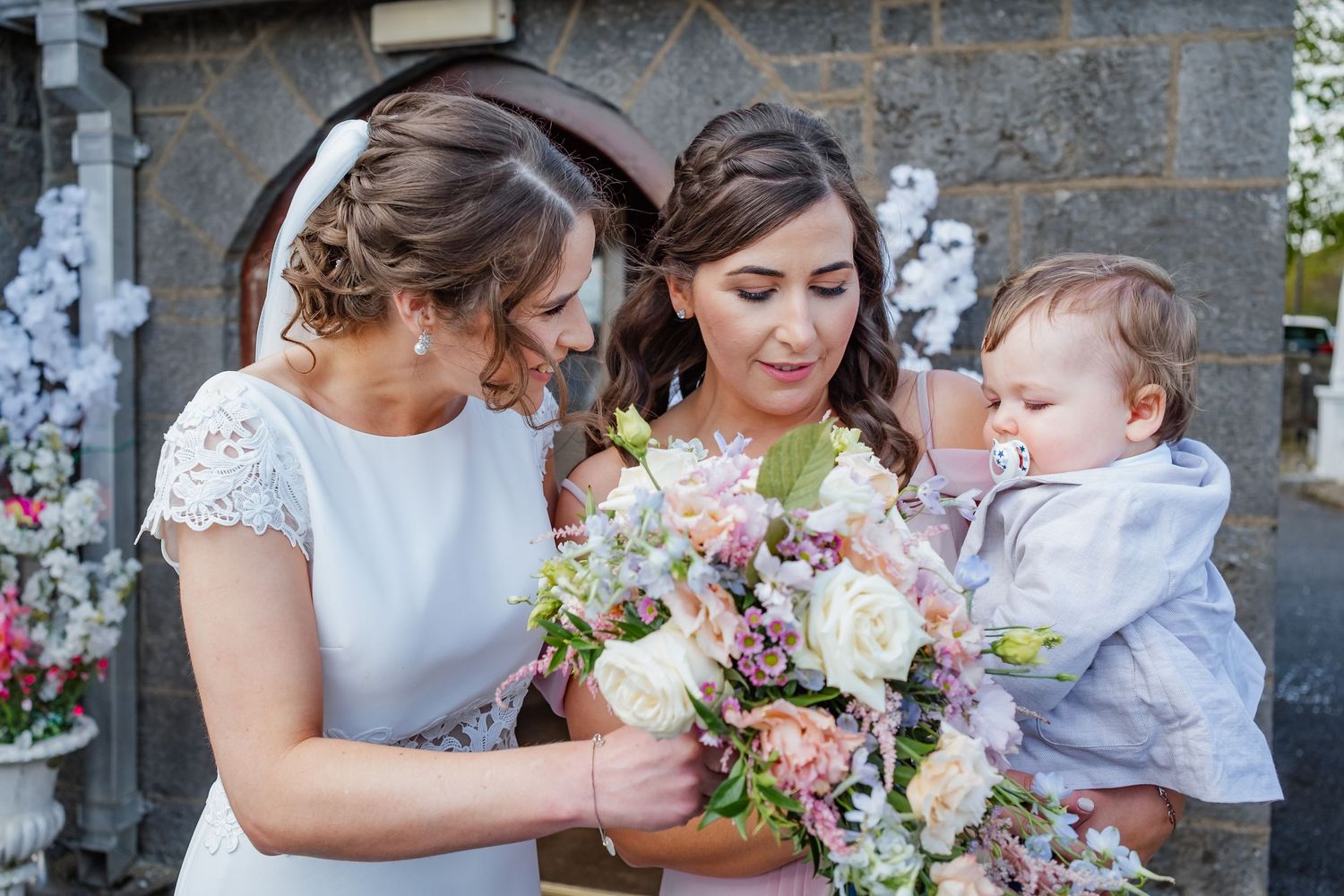 A wedding party gathers with colorful bouquets outside a stone church building.