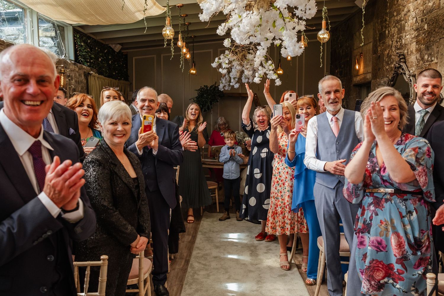 Wedding guests applaud during an elegant indoor ceremony beneath crystal chandeliers.