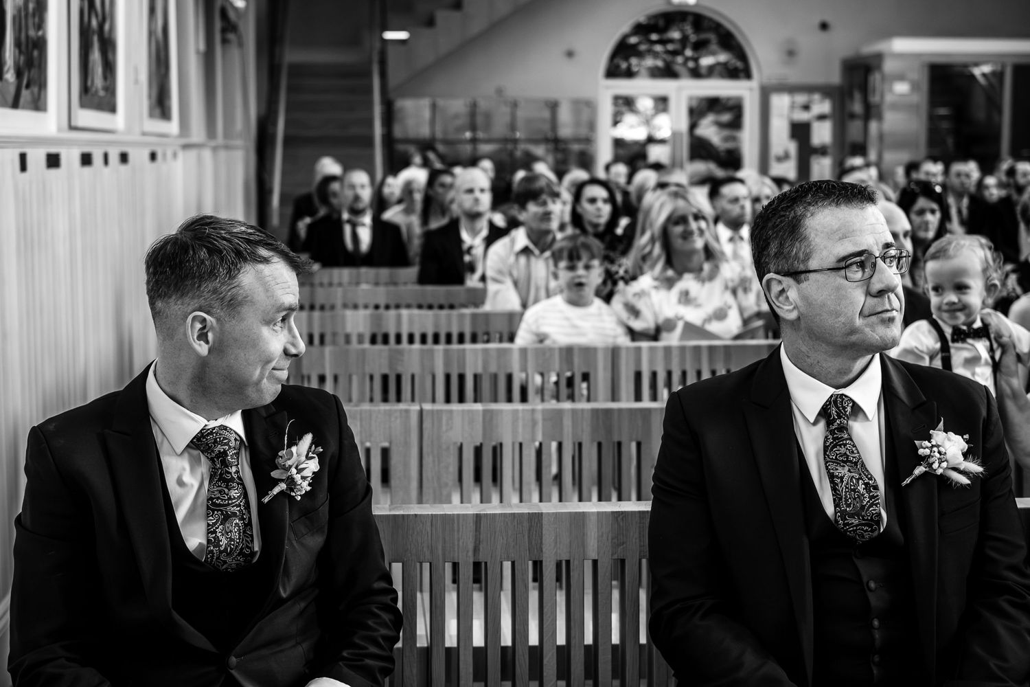 Two men in formal suits sit solemnly in the front pew during a wedding ceremony.