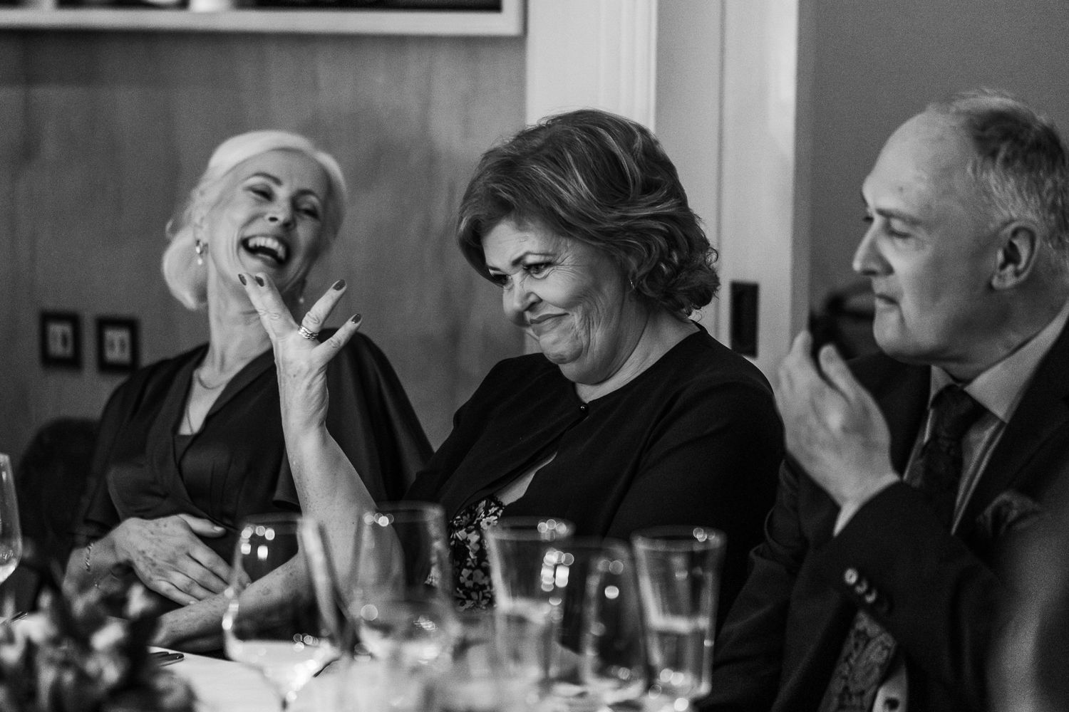 A black and white candid photo captures guests laughing heartily at a wedding reception dinner.