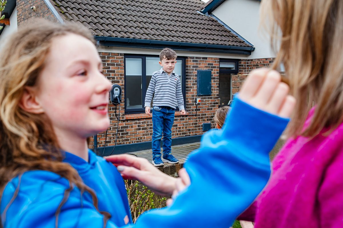 Two girls playing outside while a boy stands on a step in front of a brick house in the background.