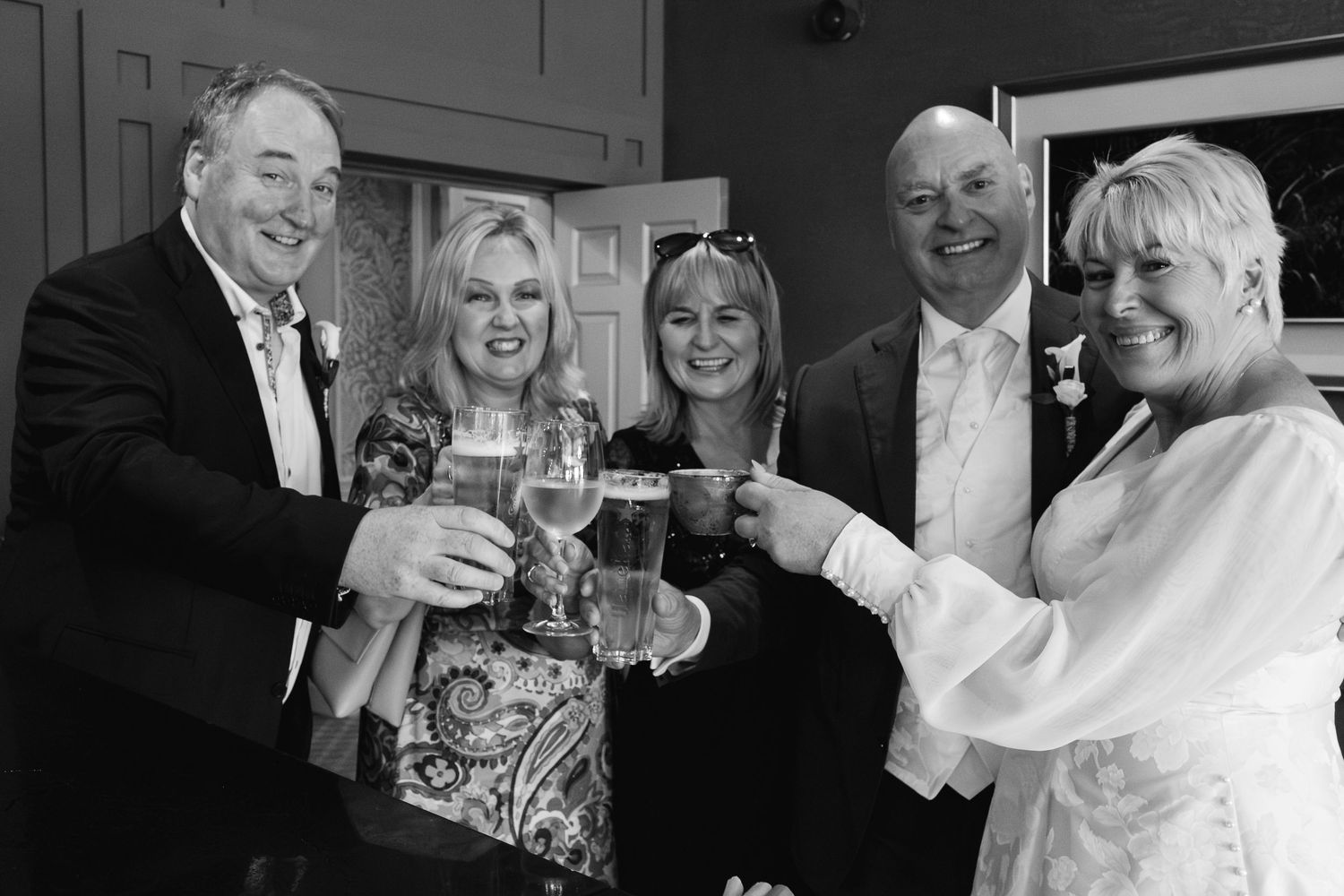 Black and white photo of five wedding guests toasting with drinks and smiling together indoors.