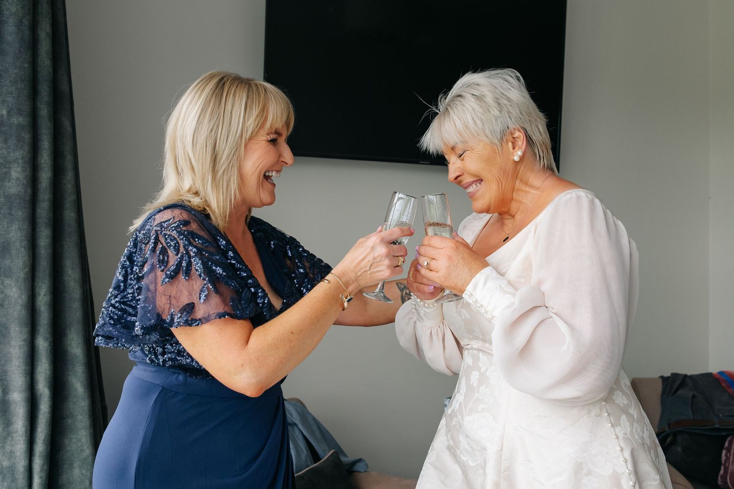 Two women in wedding attire toasting with champagne glasses during bridal preparations.
