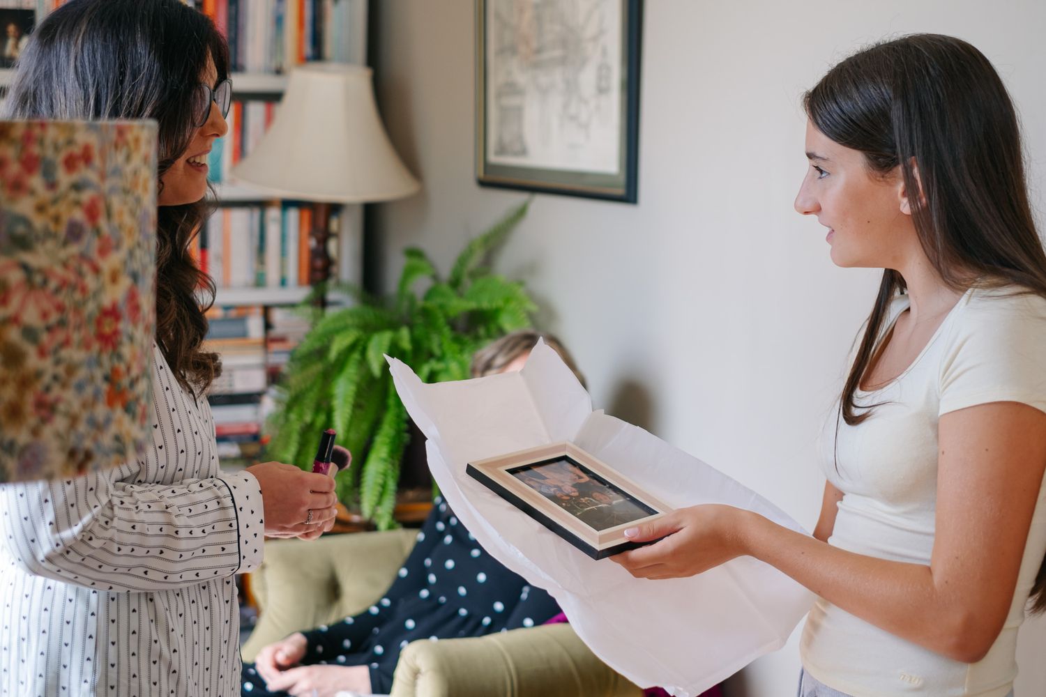 Two women examining a framed photo print together in a cozy home library setting.