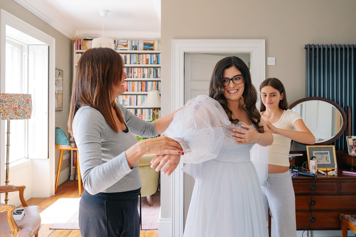 Two women help a bride in a white wedding dress get ready in a cozy home with bookshelves and a mirror.