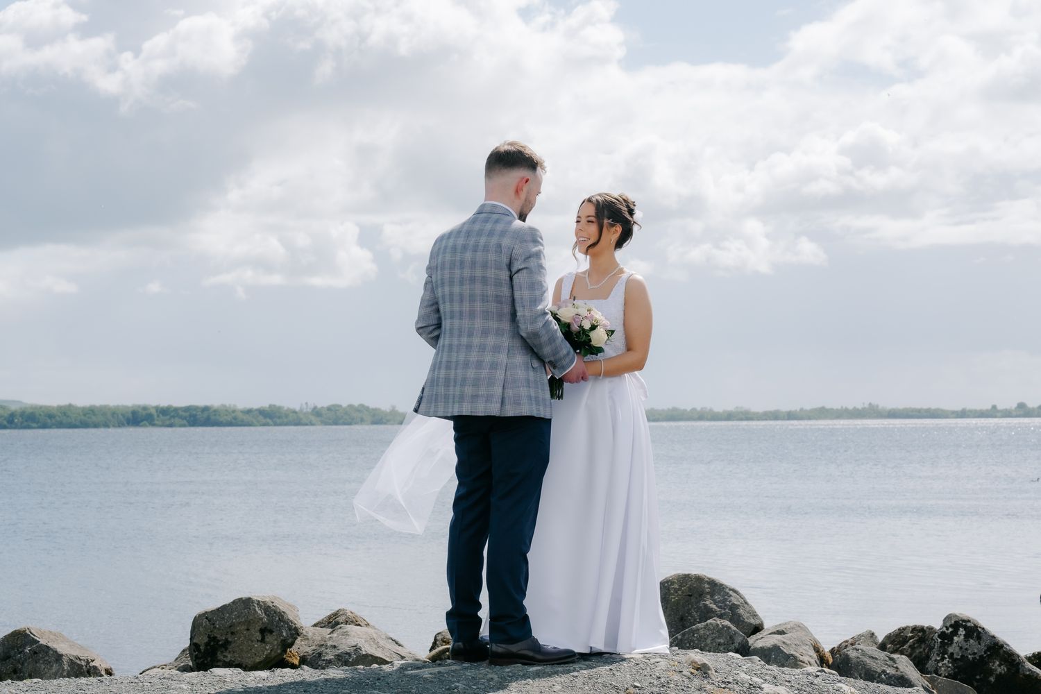 Bride and groom standing on rocks by the water, exchanging vows on their wedding day with cloudy skies.