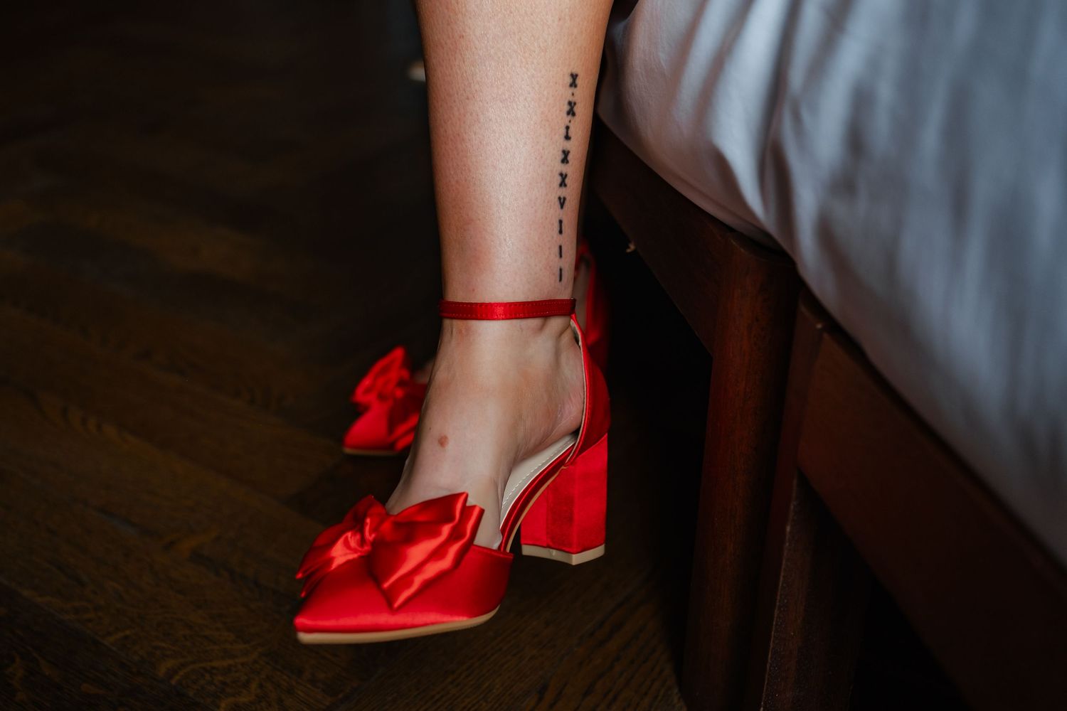 Red bow tie heels and ankle tattoo detail shot on dark wooden floor.