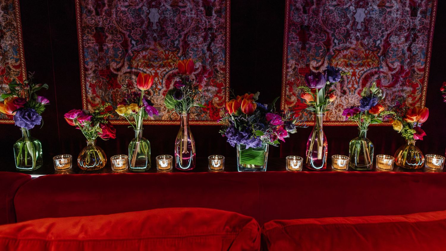 Row of colorful floral arrangements in small vases with candles against a red patterned wall.