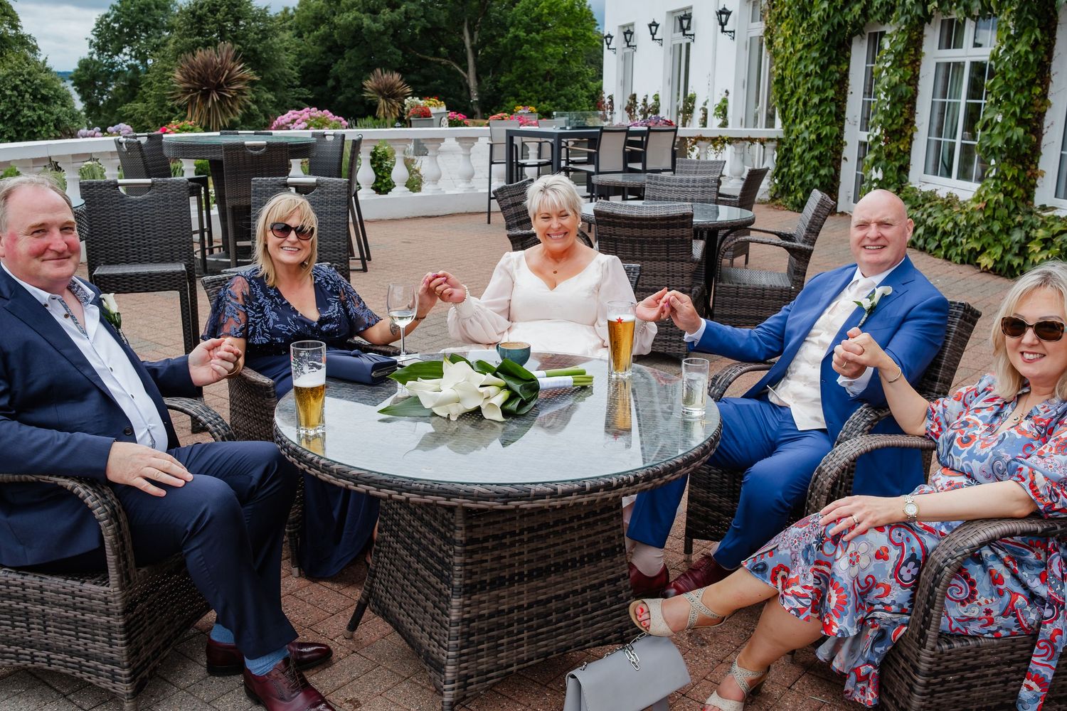 A wedding party of five guests celebrate outdoors on a patio, raising glasses with the bride and groom.
