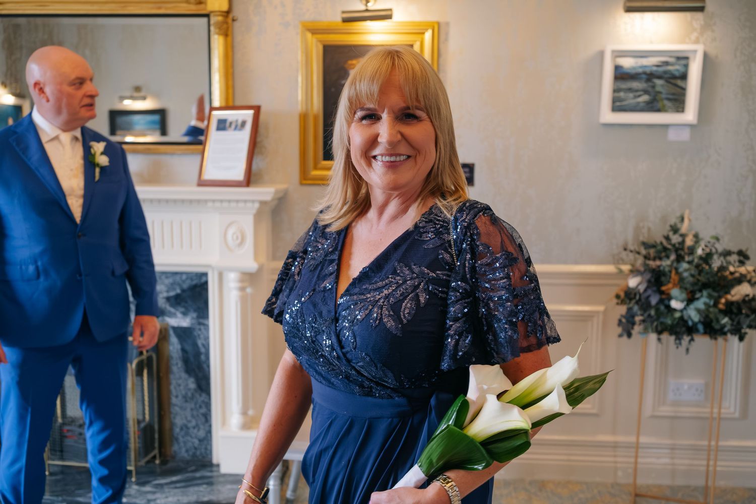 Woman in navy blue lace dress holding white calla lilies bouquet smiling at elegant wedding venue.