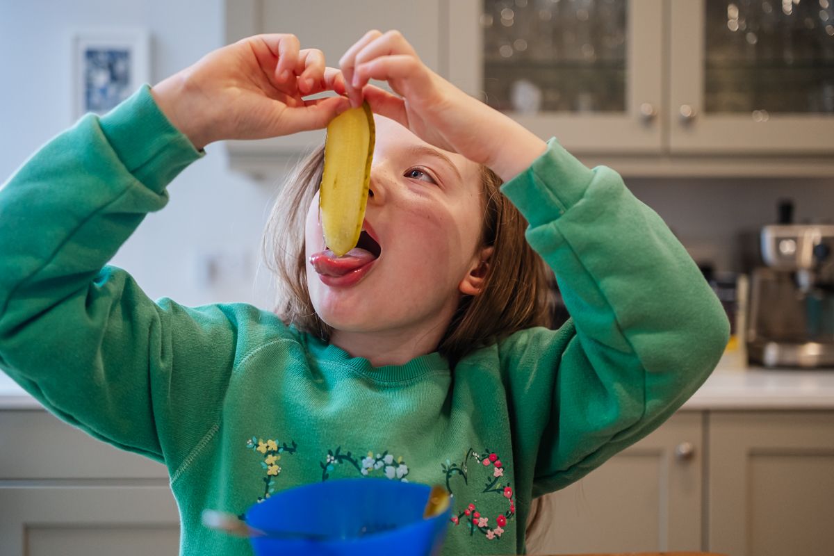 Young girl in green sweatshirt playfully holding a banana slice over her eye in a kitchen.