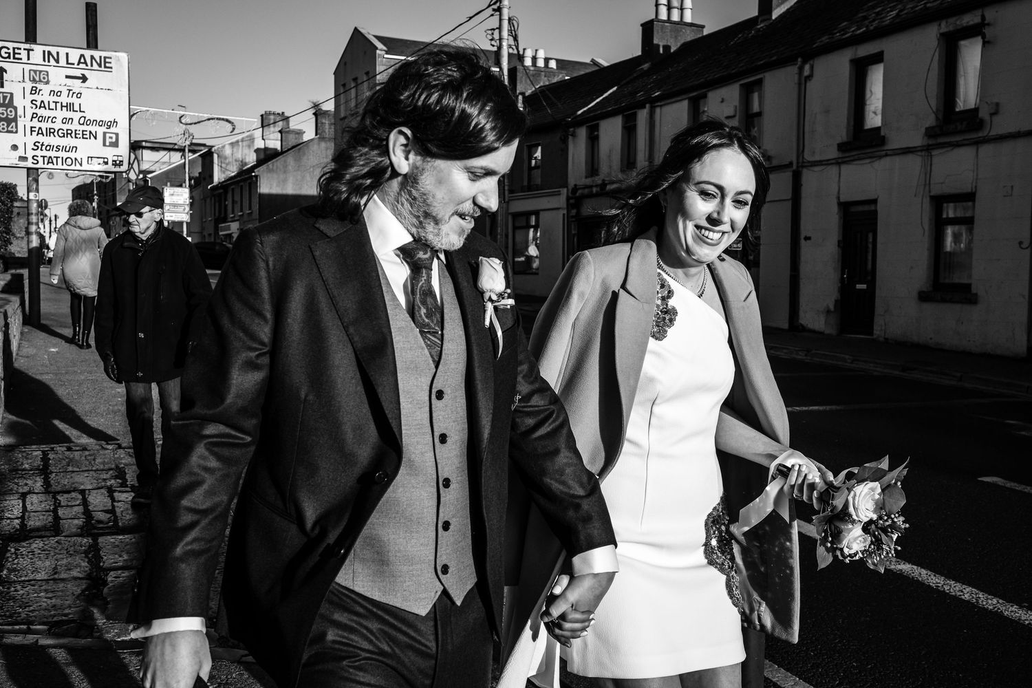 Black and white photo of a smiling bride holding flowers and groom in suit walking together on a street.