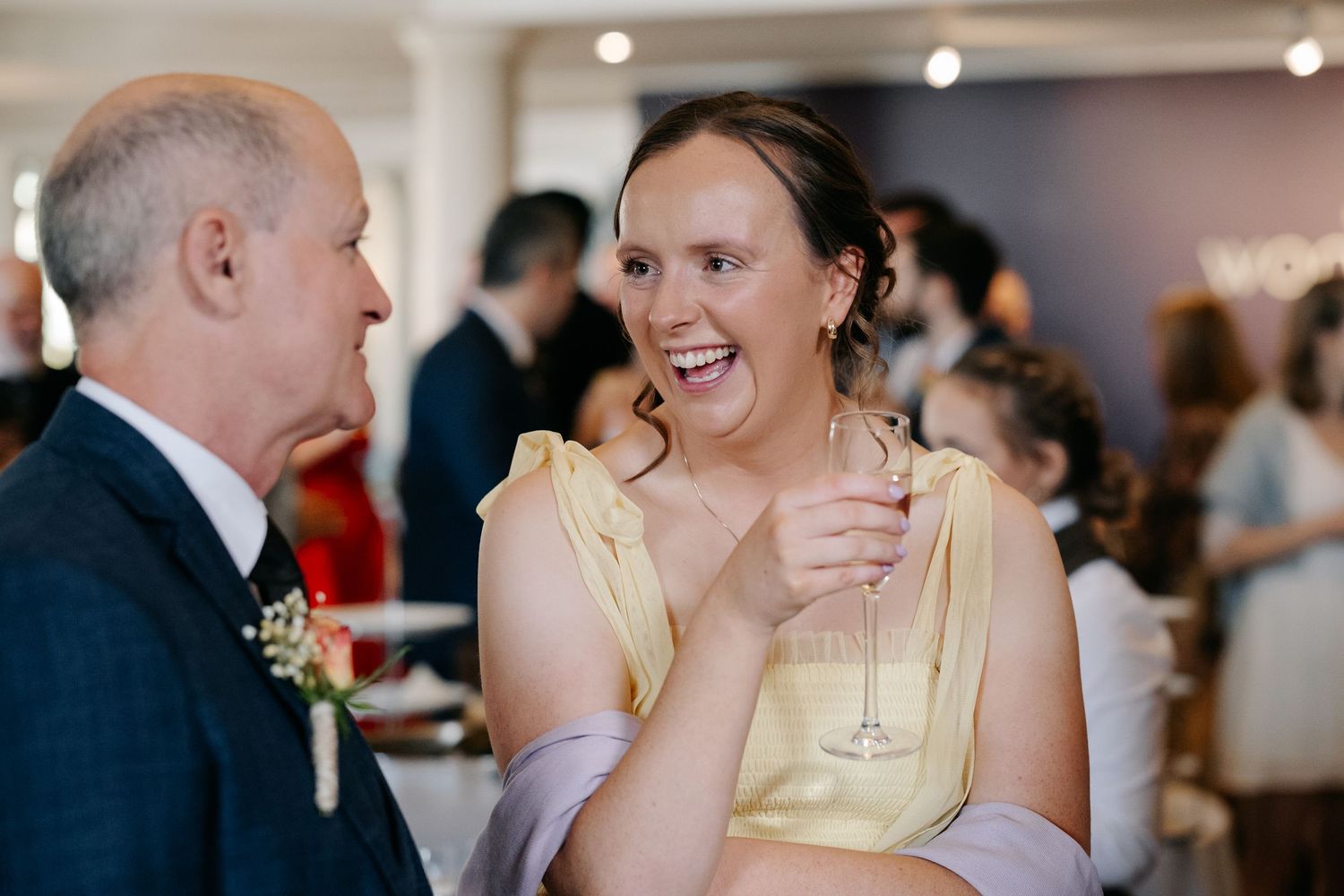 Woman in yellow dress laughing at wedding reception holding champagne glass.