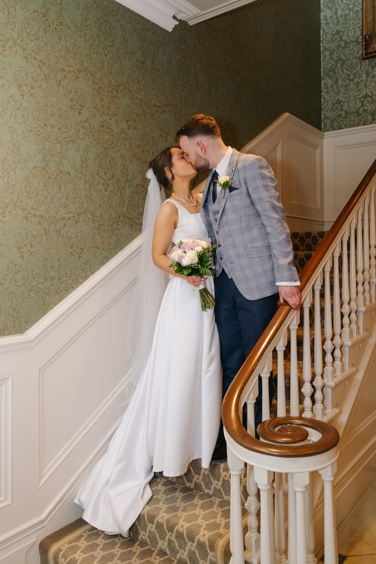 A bride and groom share a romantic kiss on an elegant staircase, she holds a bouquet in a white gown.