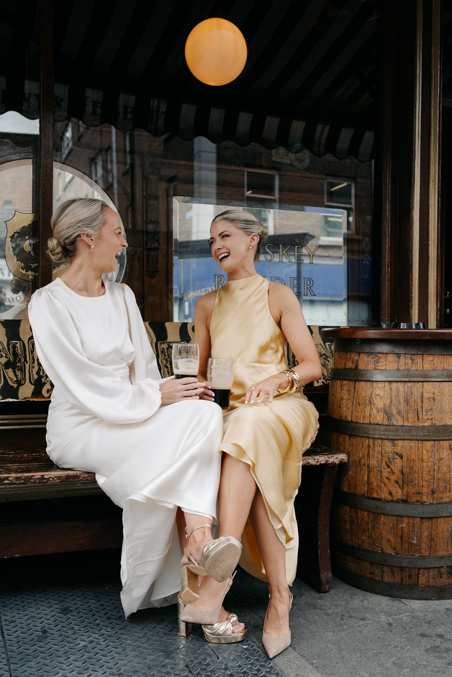 Two elegantly dressed women laughing together outside a pub, one in white and one in gold satin dress.