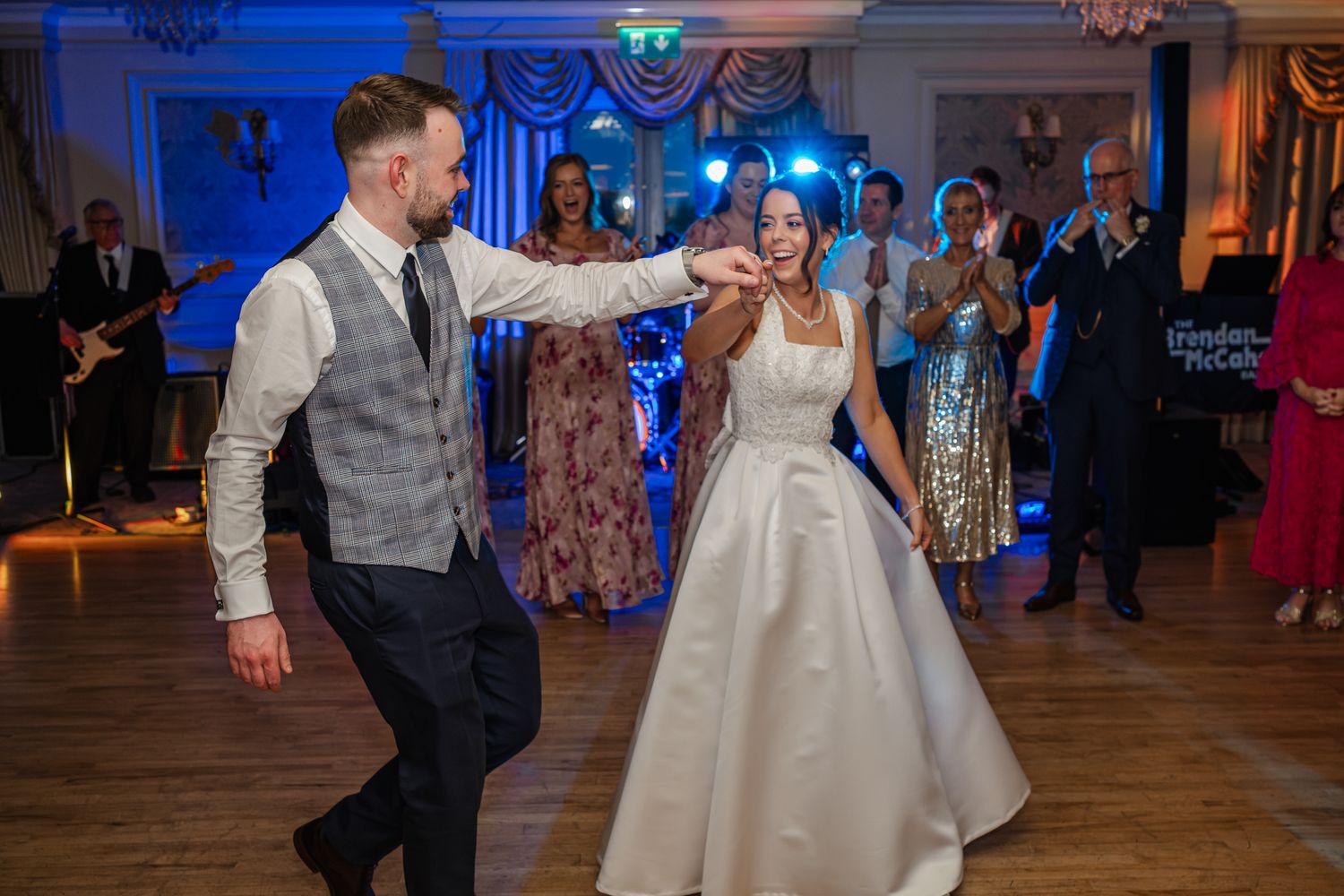 Bride and groom share their first dance at elegant wedding reception with live band and guests applauding in background.