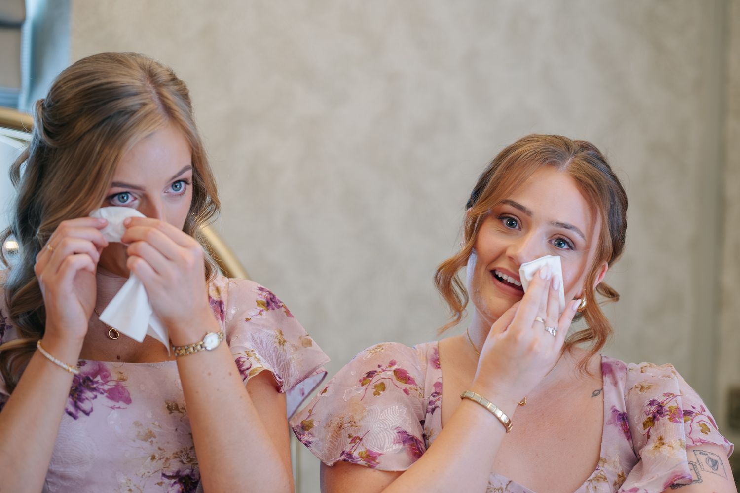 Two bridesmaids in floral dresses wiping tears with tissues during an emotional wedding preparation moment.