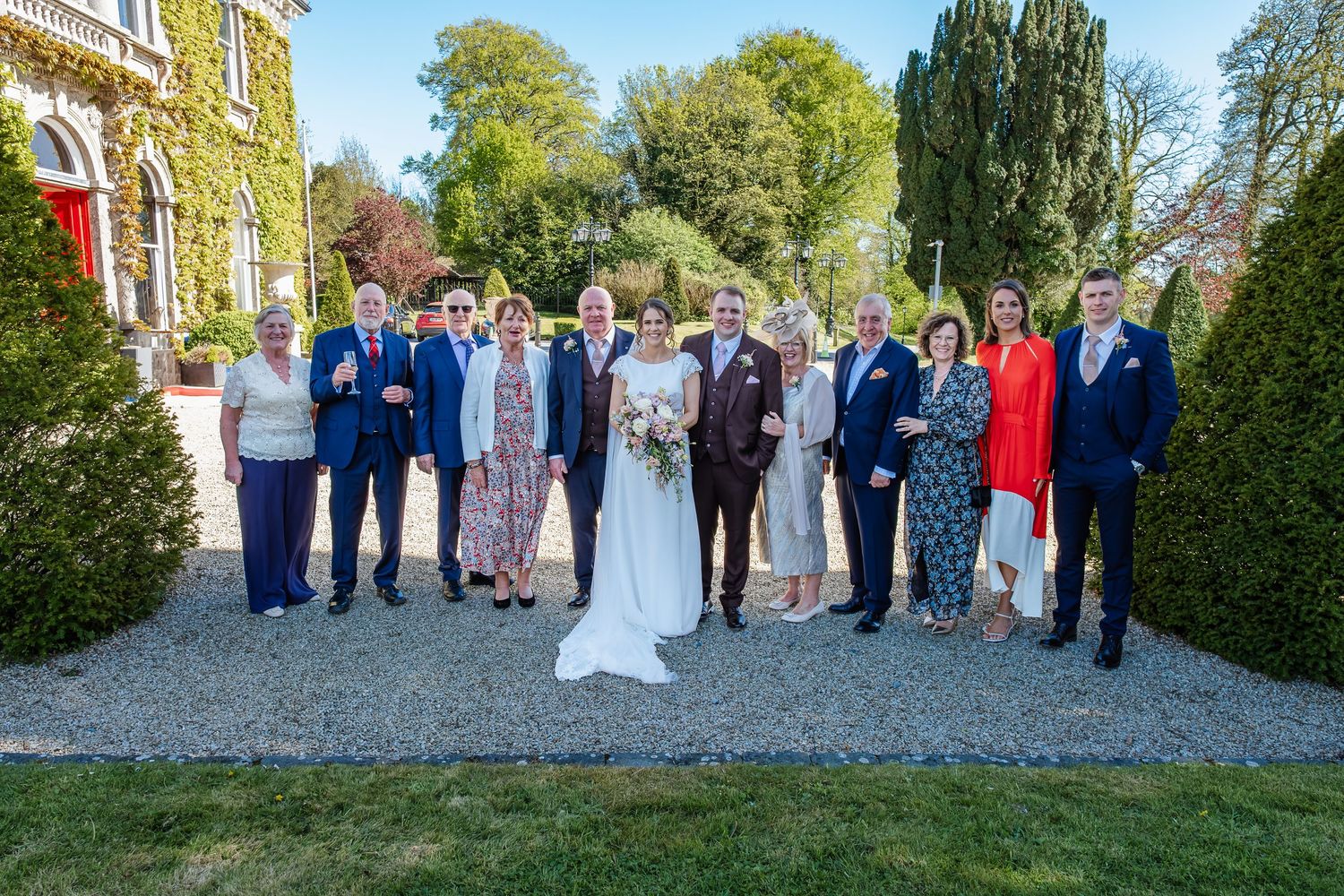 Wedding party group posing together in front of ivy-covered manor house on sunny garden grounds.