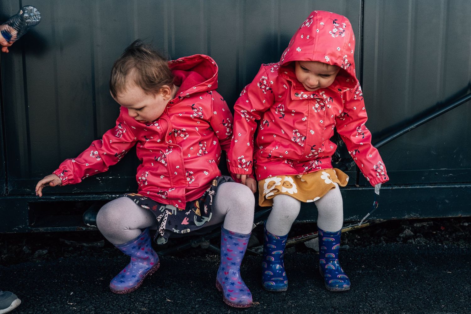 Two children in matching red patterned jackets and rain boots sit on a bench against a dark wall.
