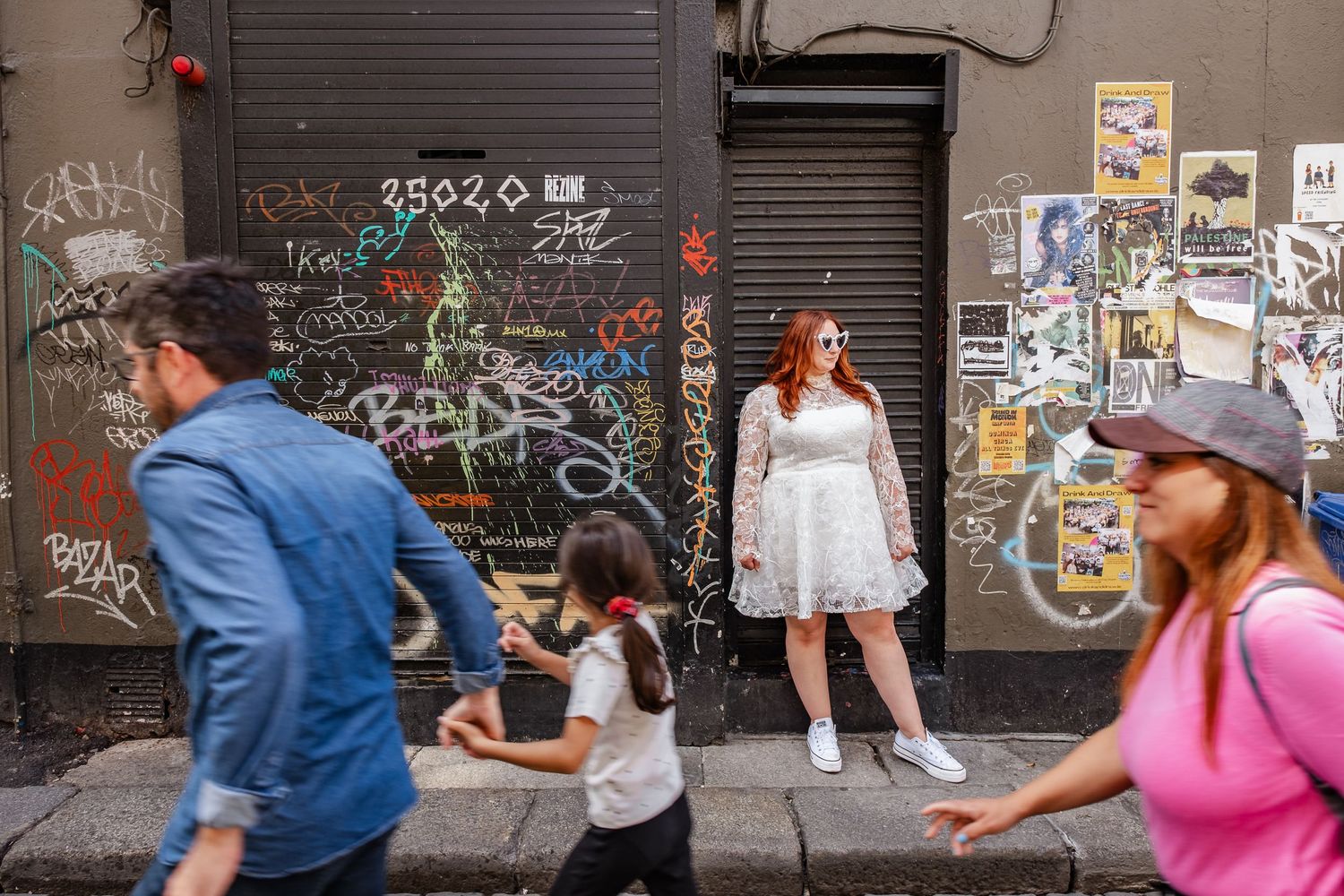 Street scene featuring people walking past graffiti covered walls and posters in an urban setting.