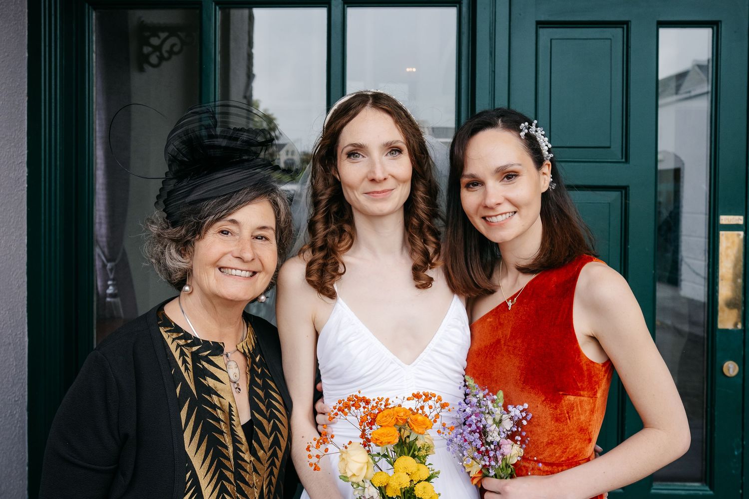 Wedding party group photo with bridesmaids in colorful dresses holding bouquets against green doors.