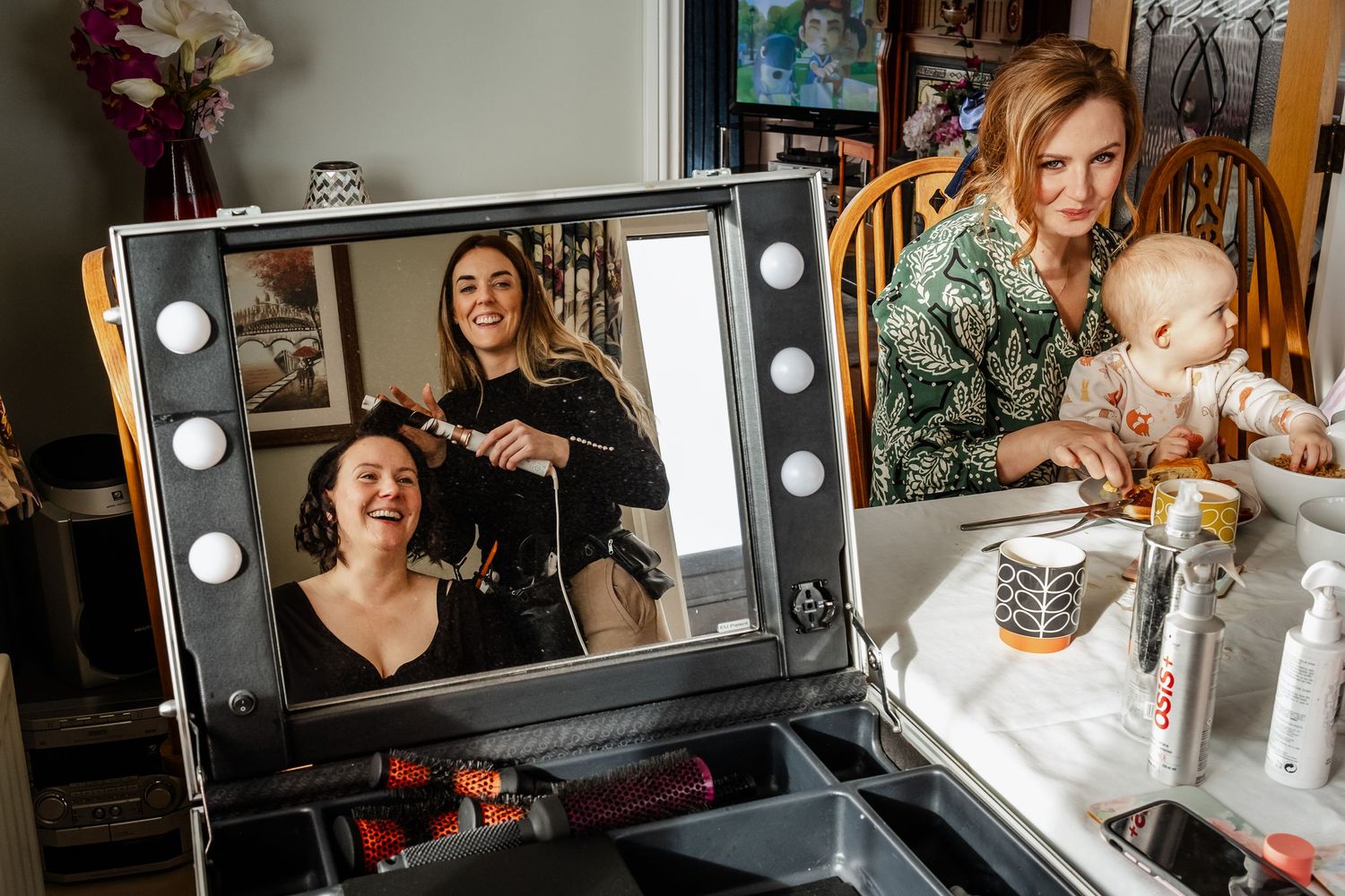 A hairstylist styling a woman's hair reflected in a lighted vanity mirror, while another woman holds a baby nearby.