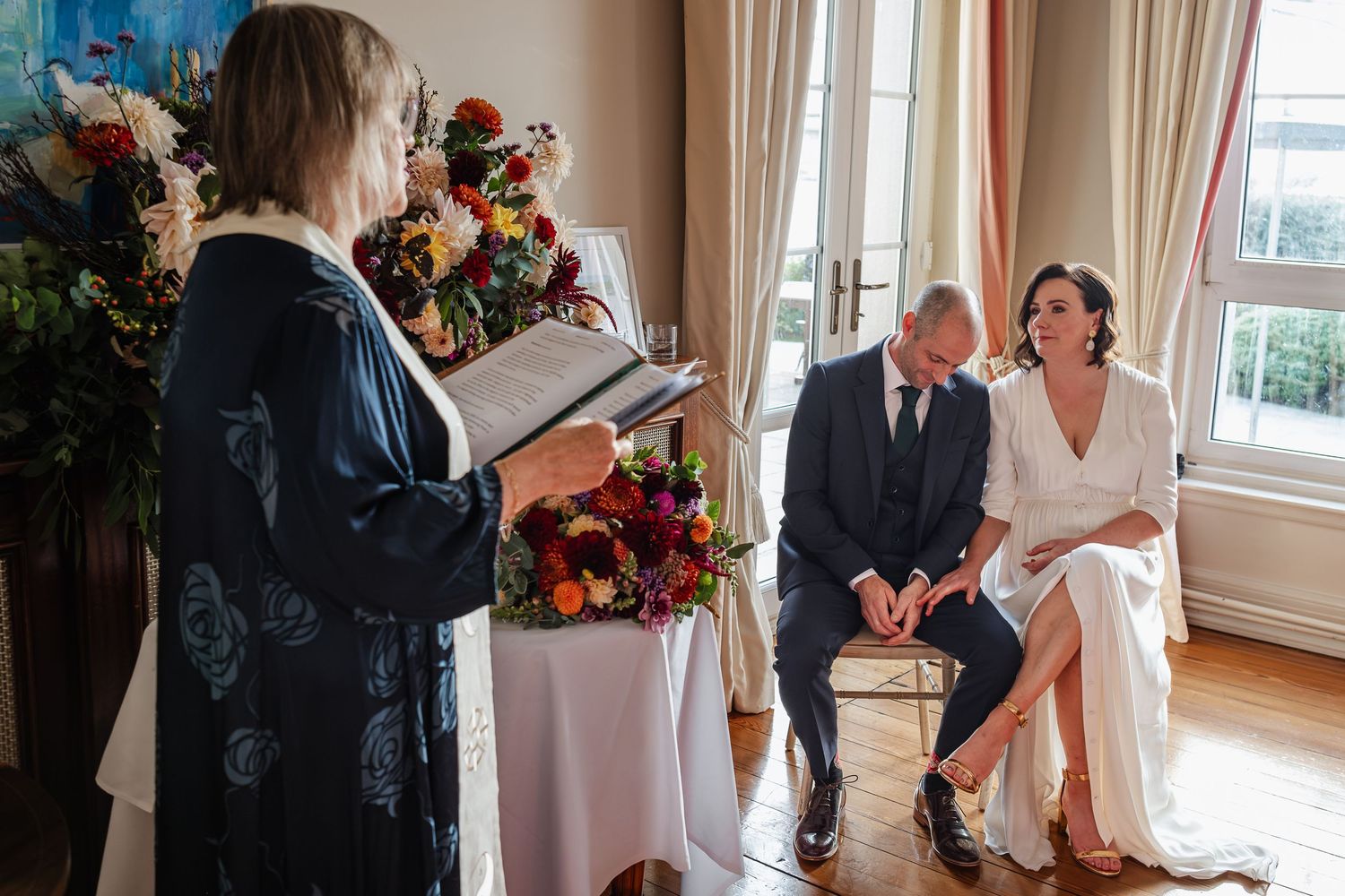 A couple sits together during an intimate wedding ceremony while an officiant reads from a book beside floral arrangements.
