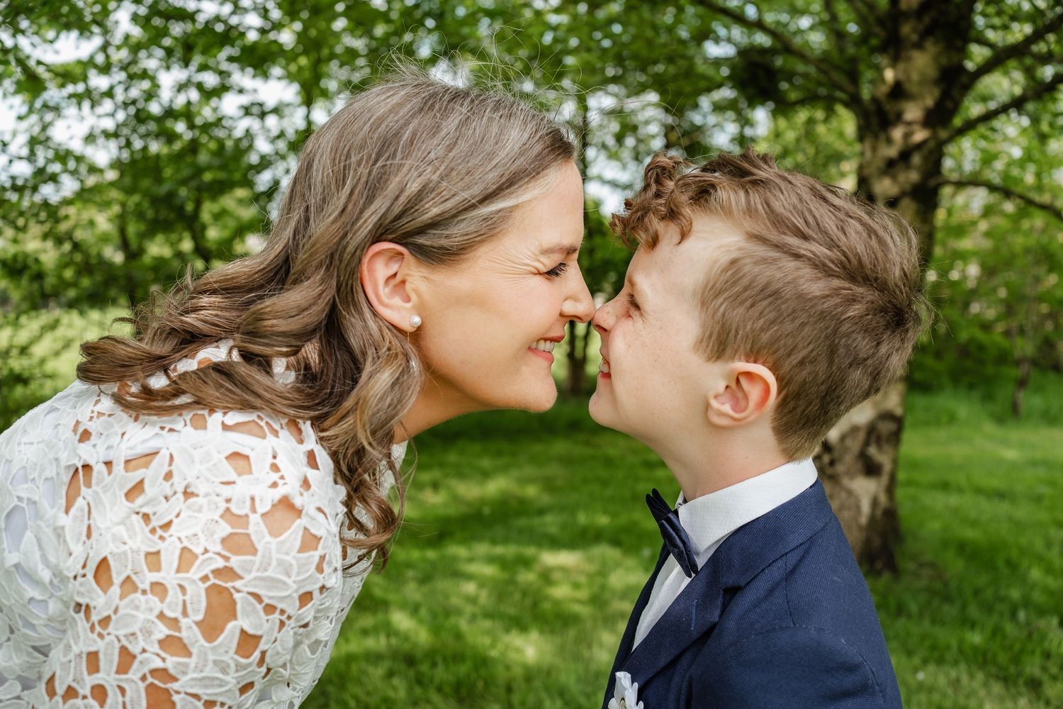 Bride in white lace dress shares a tender nose-to-nose moment with young boy in navy suit outdoors.