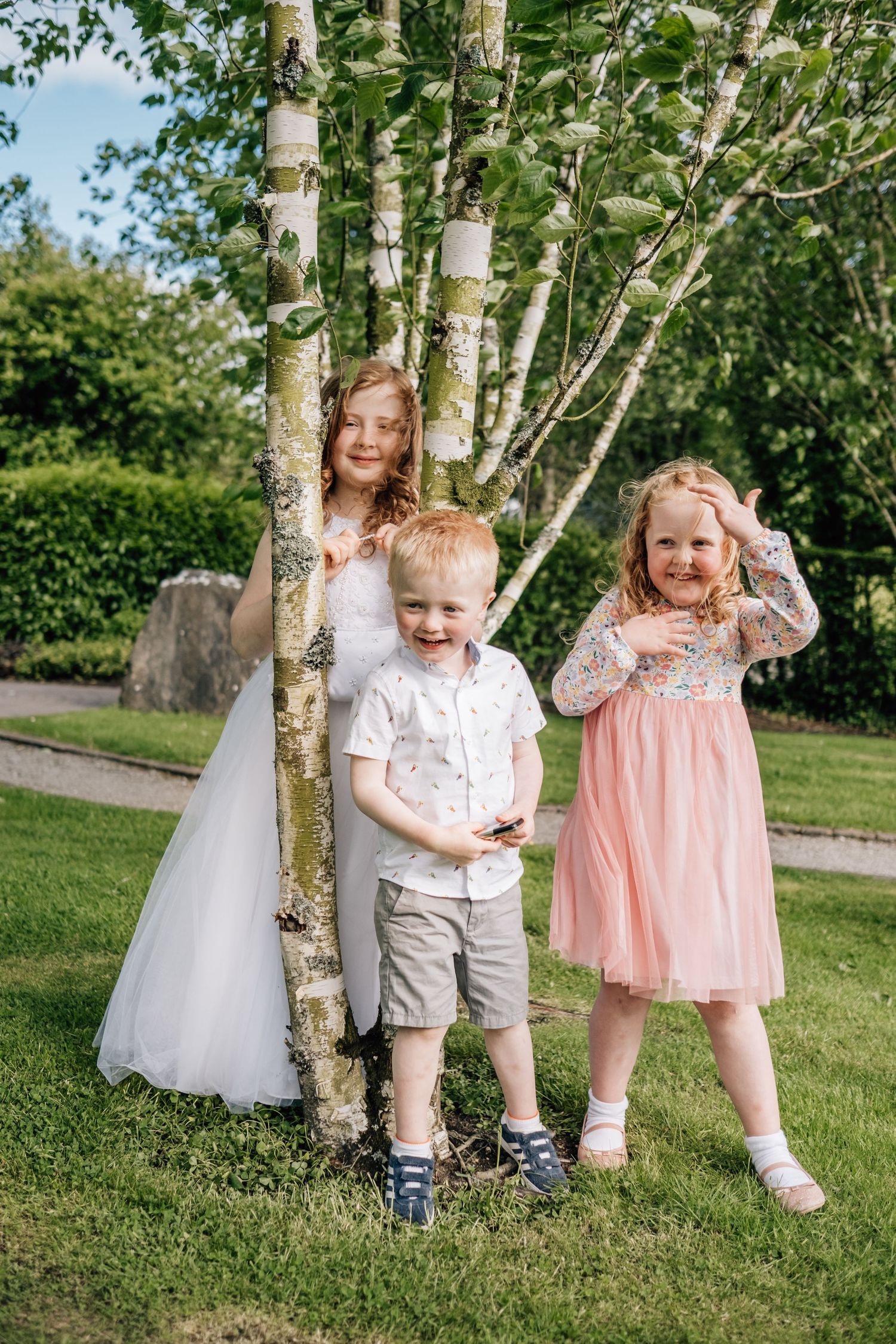 Three children posing outdoors near birch trees, wearing formal attire including white dress, pink tulle dress, and dress shirt.