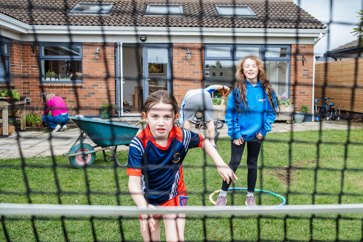 Two children play tennis in a backyard garden, viewed through the net on a cloudy day.